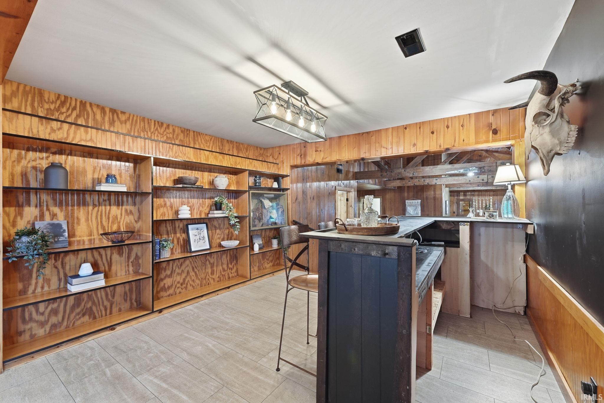 Built-in wood shelving, wood paneling, L-shaped bar with dark wood finish, light tile flooring, and a multi-bulb chandelier