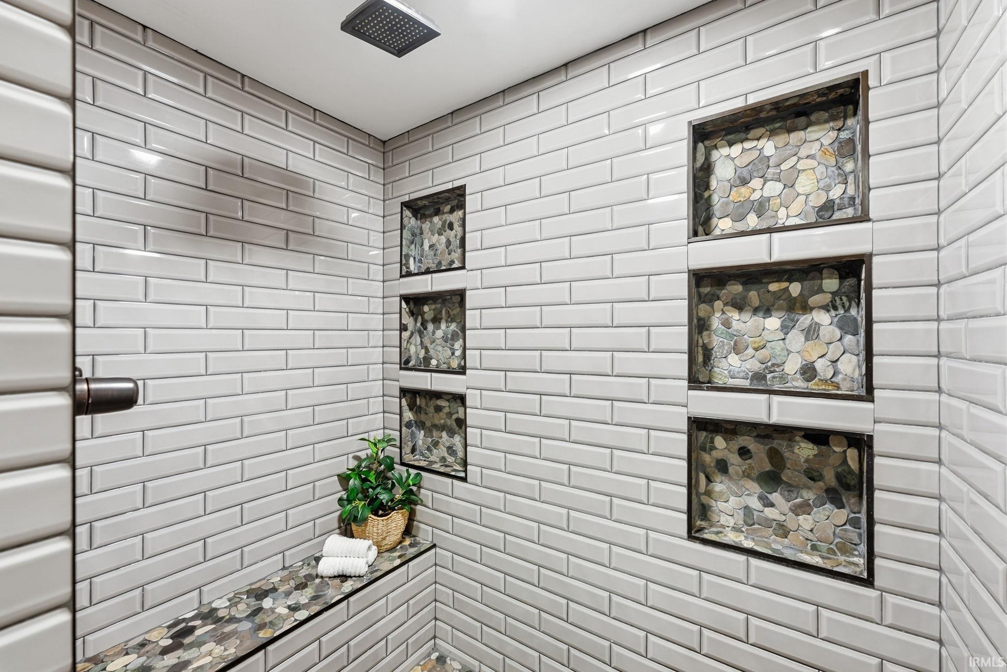 White subway tile surround with pebble tile accents, built-in shelving, a rainfall showerhead, and a fixed shower bench