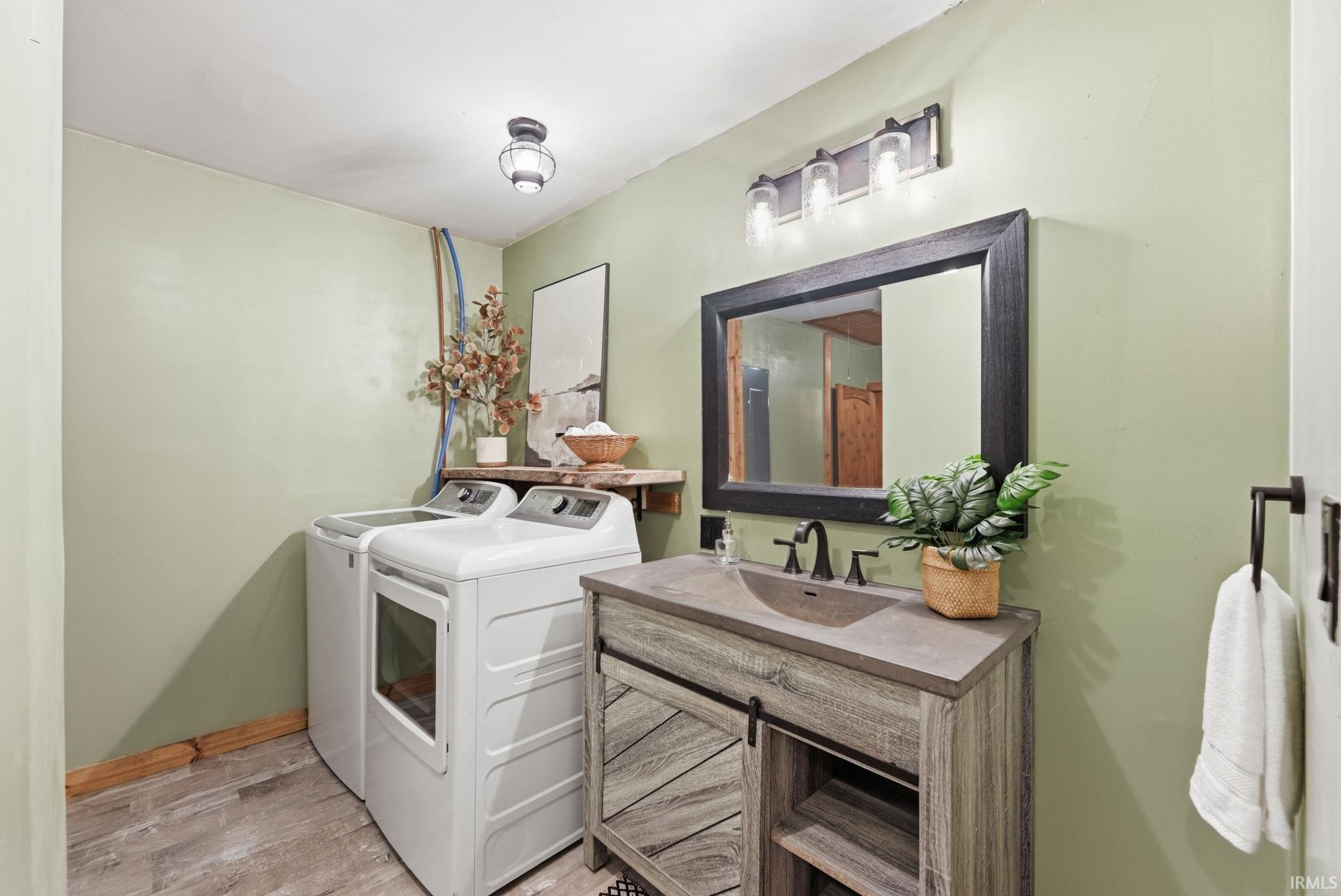Laundry area with wood-finish flooring, a vanity with a dark gray countertop, black framed mirror, and a wall-mounted light fixture