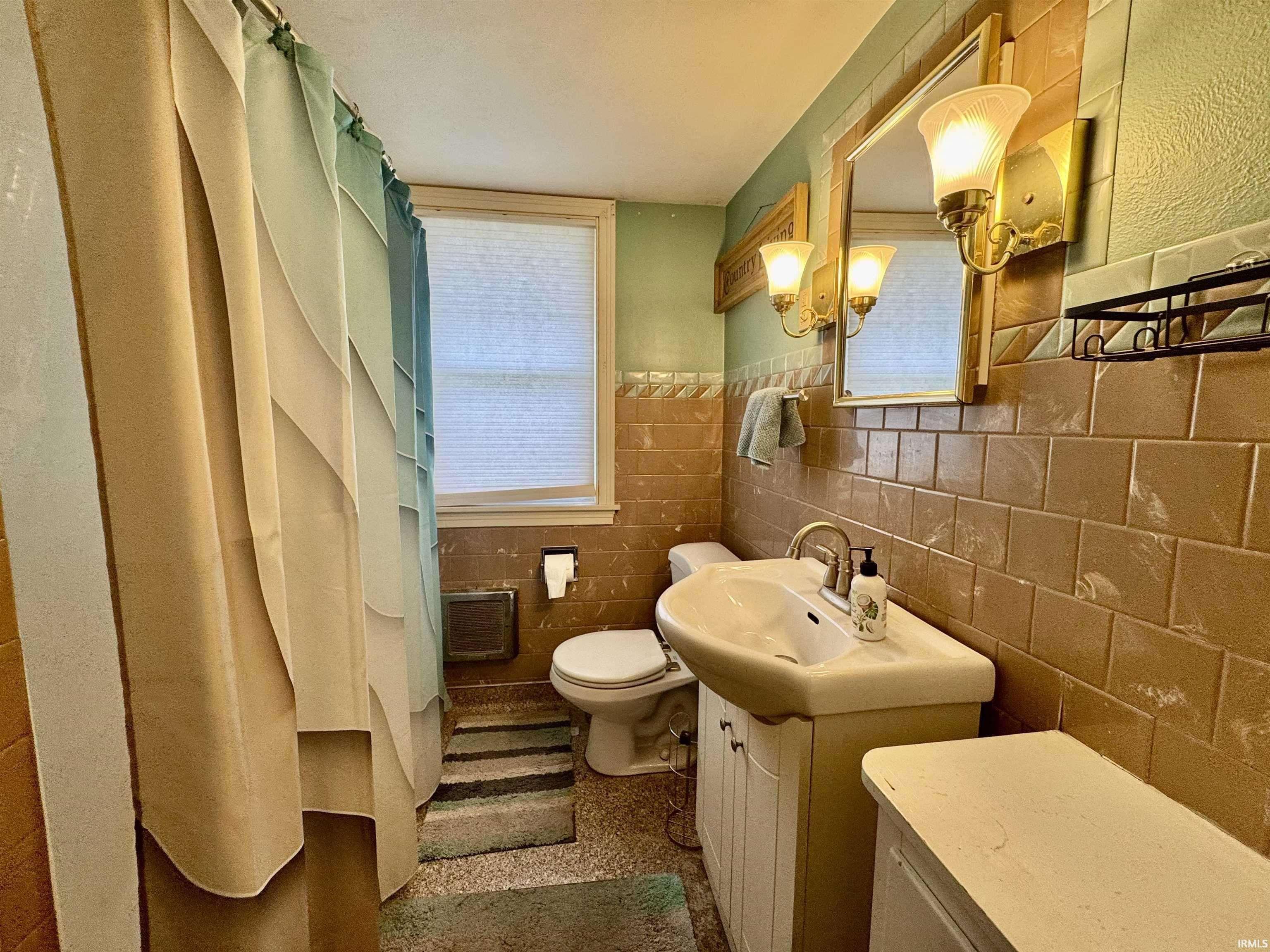 Bathroom featuring tiled walls, pedestal sink, wall-mounted vanity lighting, and a window with blinds