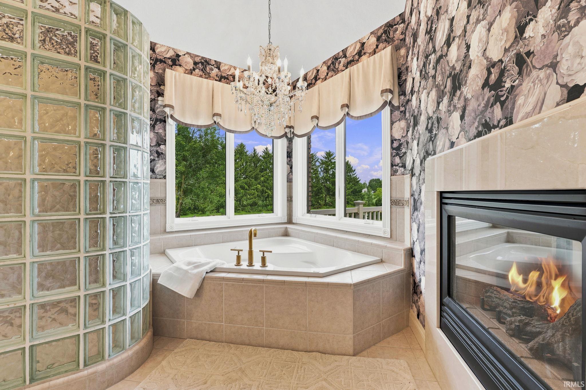 Luxurious primary bath featuring dual vanities, furniture-style custom cabinetry, glass block walk-in shower, and soaking tub with pond views and fireplace.