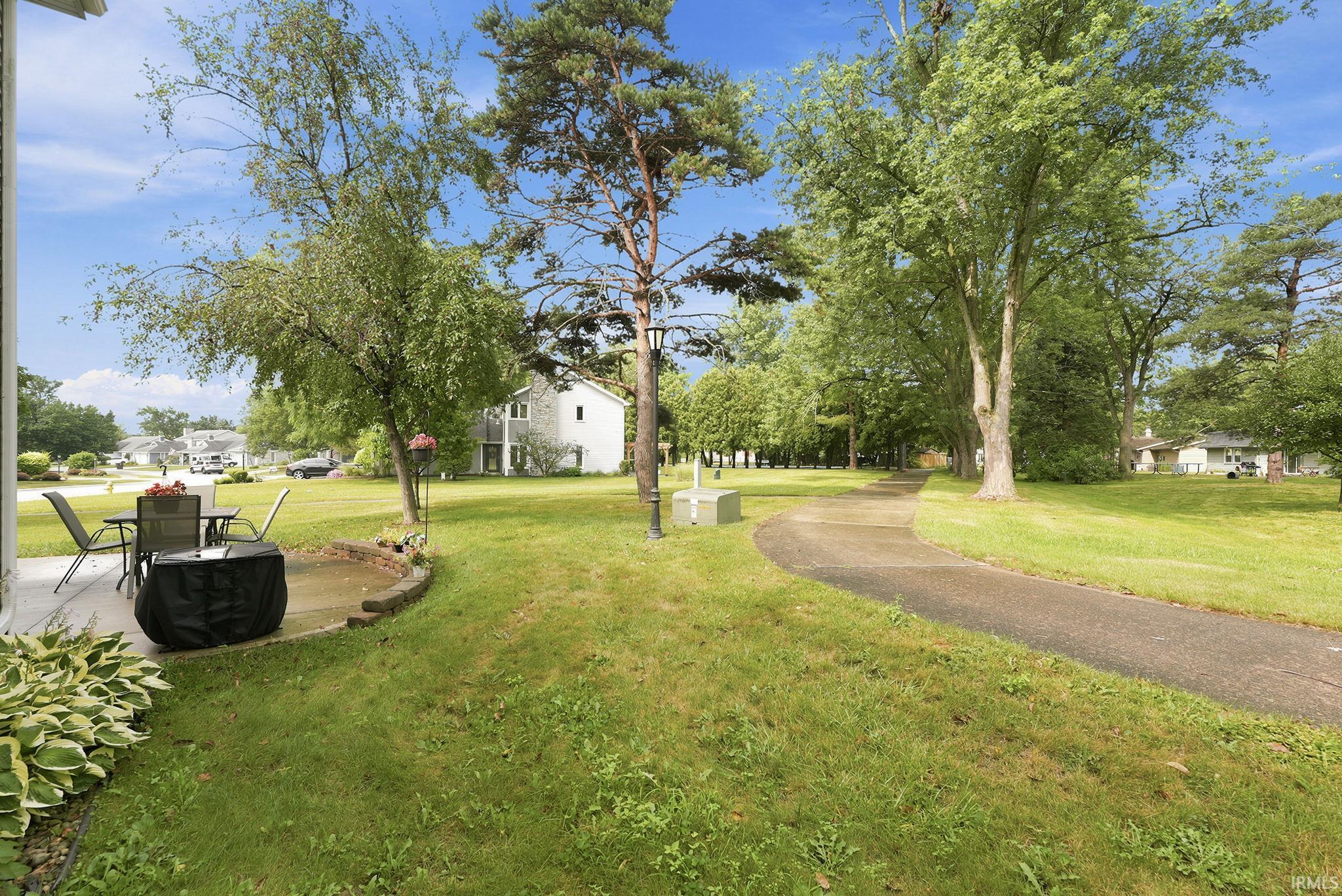 Expansive green space featuring a paved pathway, mature trees, and a concrete patio area with a low retaining wall