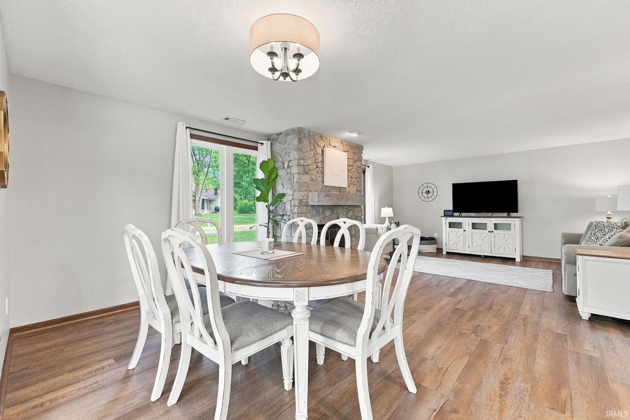 Open-concept dining area with wood-finish flooring, a contemporary drum chandelier, and a stone-clad fireplace visible in the background