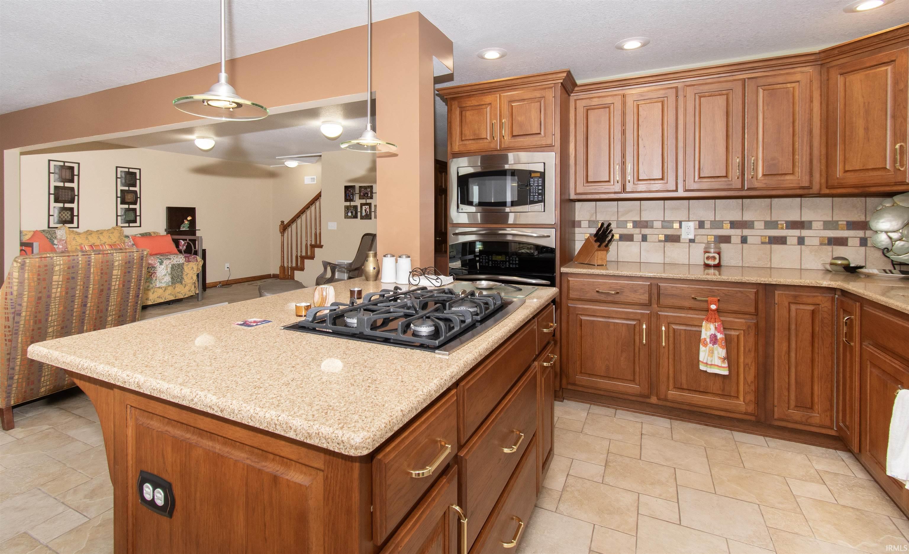Kitchen featuring an island with a built-in gas cooktop, pendant lighting, warm wood cabinetry, neutral tile backsplash, and integrated stainless steel appliances