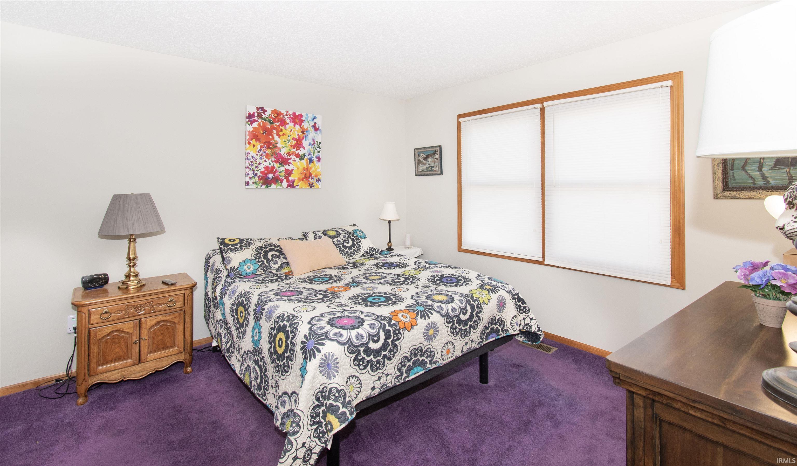 Bedroom featuring neutral walls, purple carpeting, and a double window with light wood trim and white roller blinds