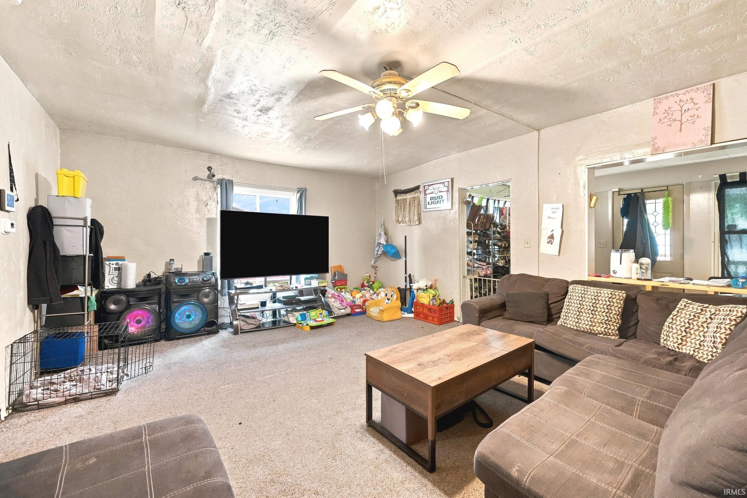 Carpeted living area featuring a ceiling fan with light fixture, a window with vertical blinds, and a built-in pass-through opening