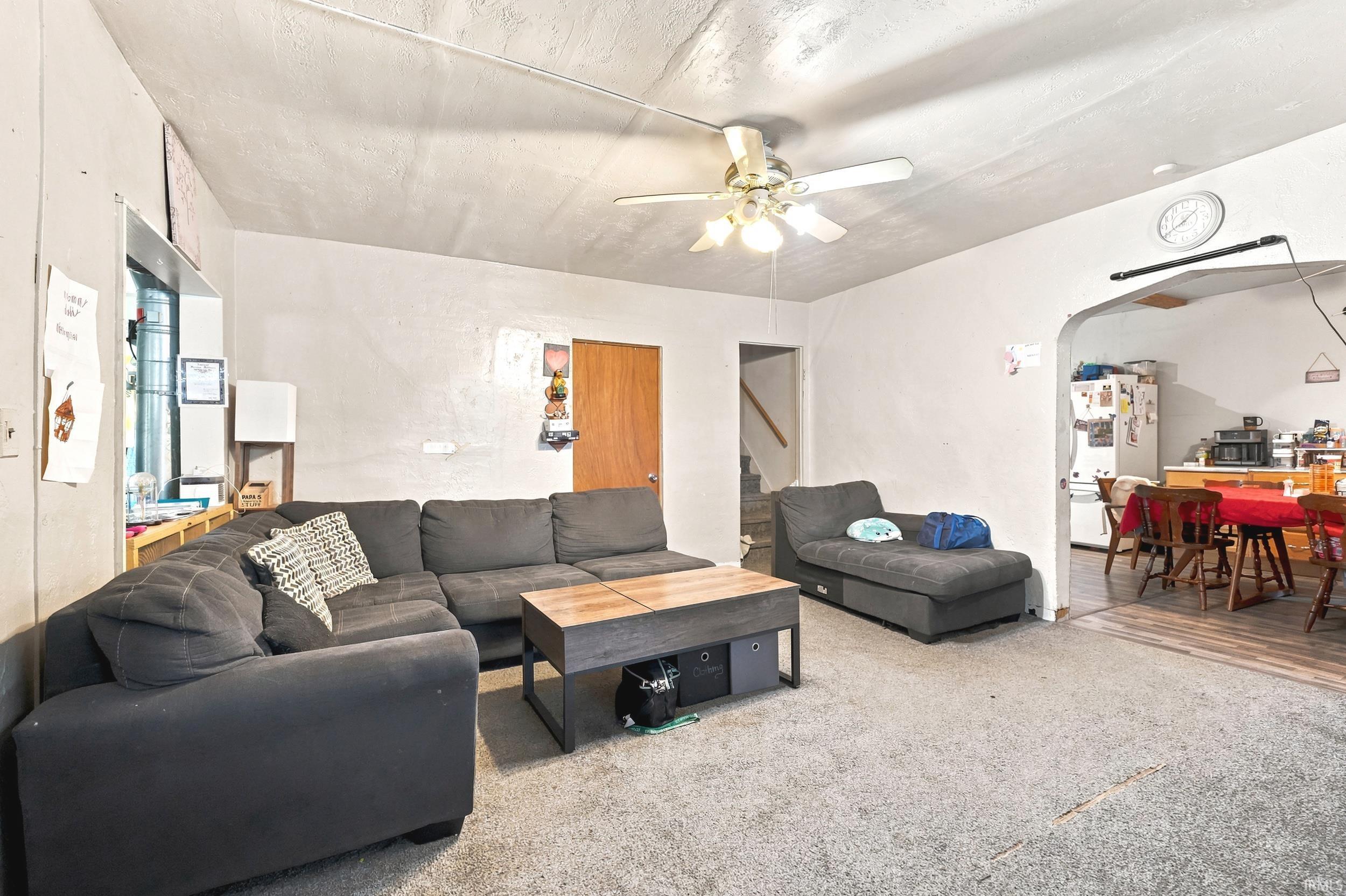Carpeted living area featuring a ceiling fan, a window with natural light, and an arched doorway opening to a dining space with wood-finish flooring