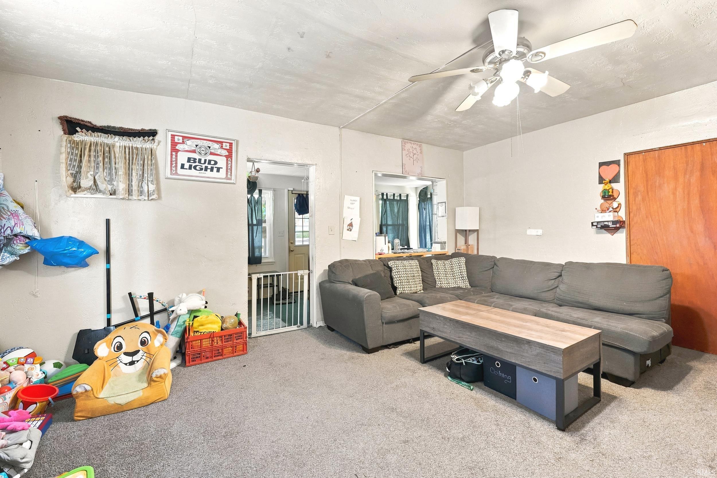 Carpeted living area featuring a ceiling fan with light fixture, a wood-finish door, and doorway openings to additional spaces