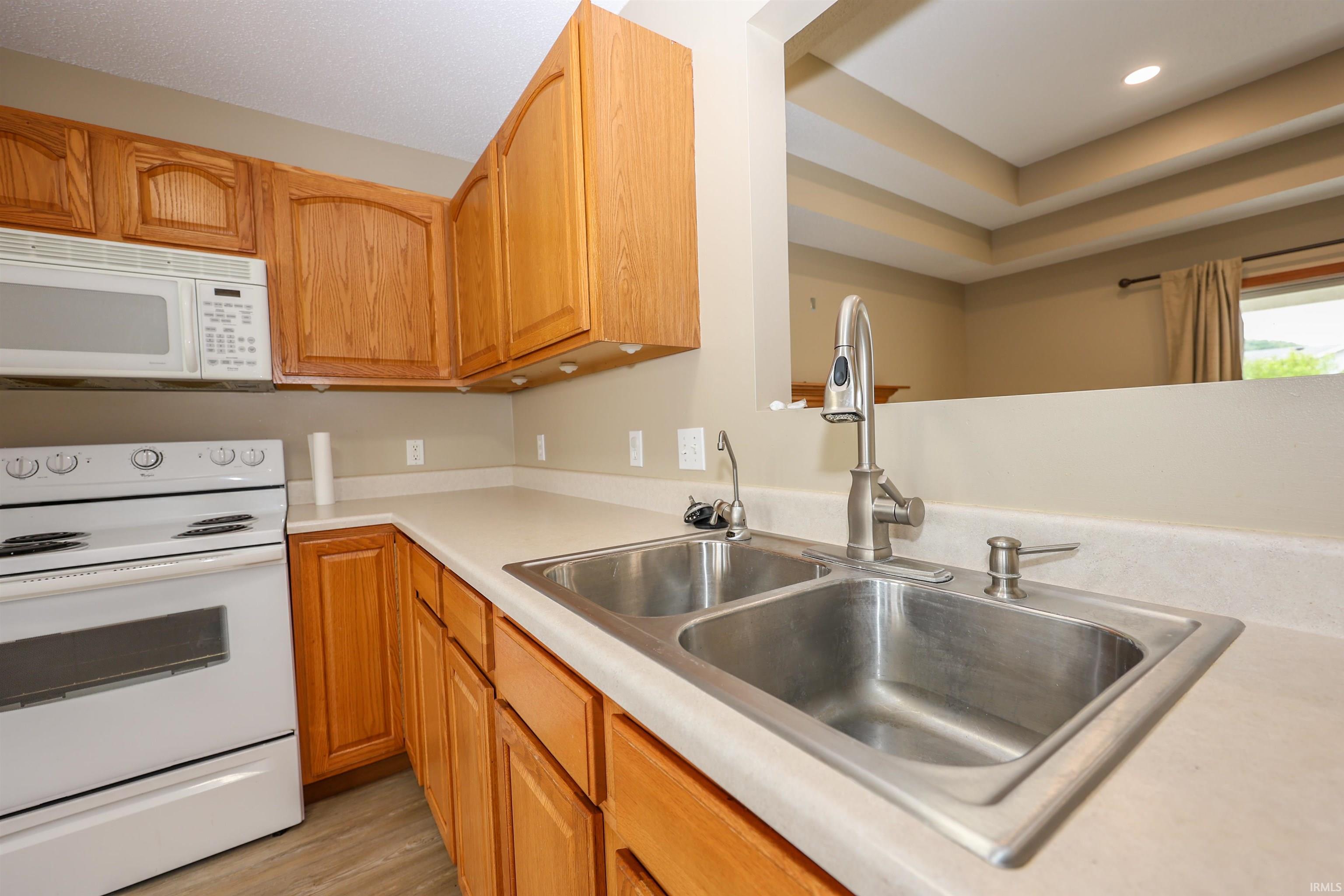 Kitchen featuring wood cabinetry, a double-basin stainless steel sink with a brushed nickel faucet, light-toned countertops, white range, and a white over-the-range microwave