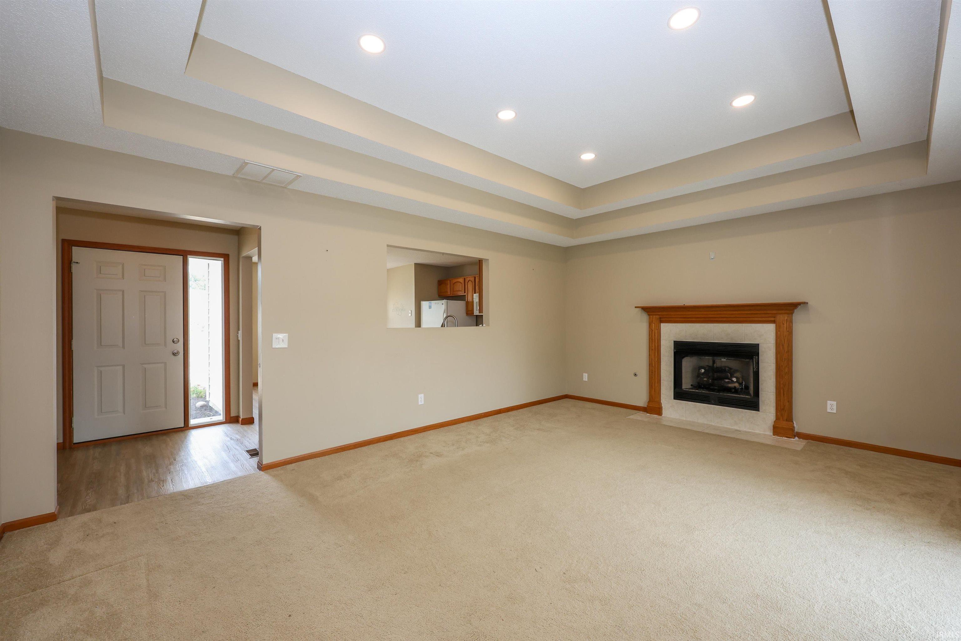 Spacious living area featuring a tray ceiling with recessed lighting, an integrated fireplace with a wood mantel and stone surround, and plush carpet flooring