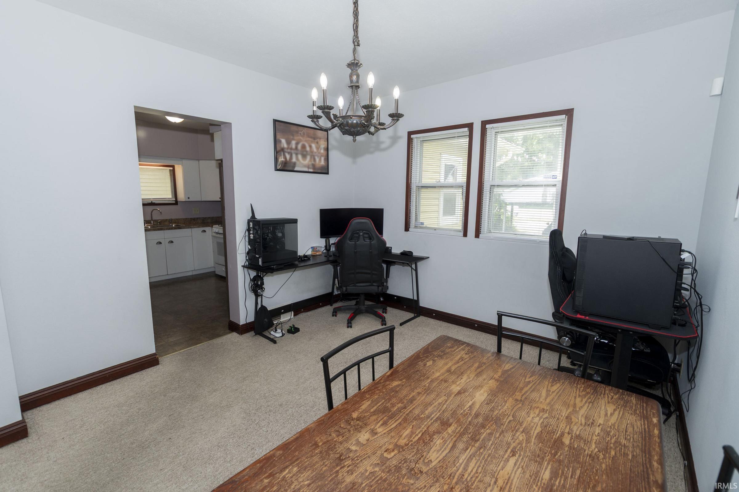 Carpeted room with a dark-finished chandelier, two windows, and a doorway opening to a kitchen featuring white cabinetry and a stone-finish countertop