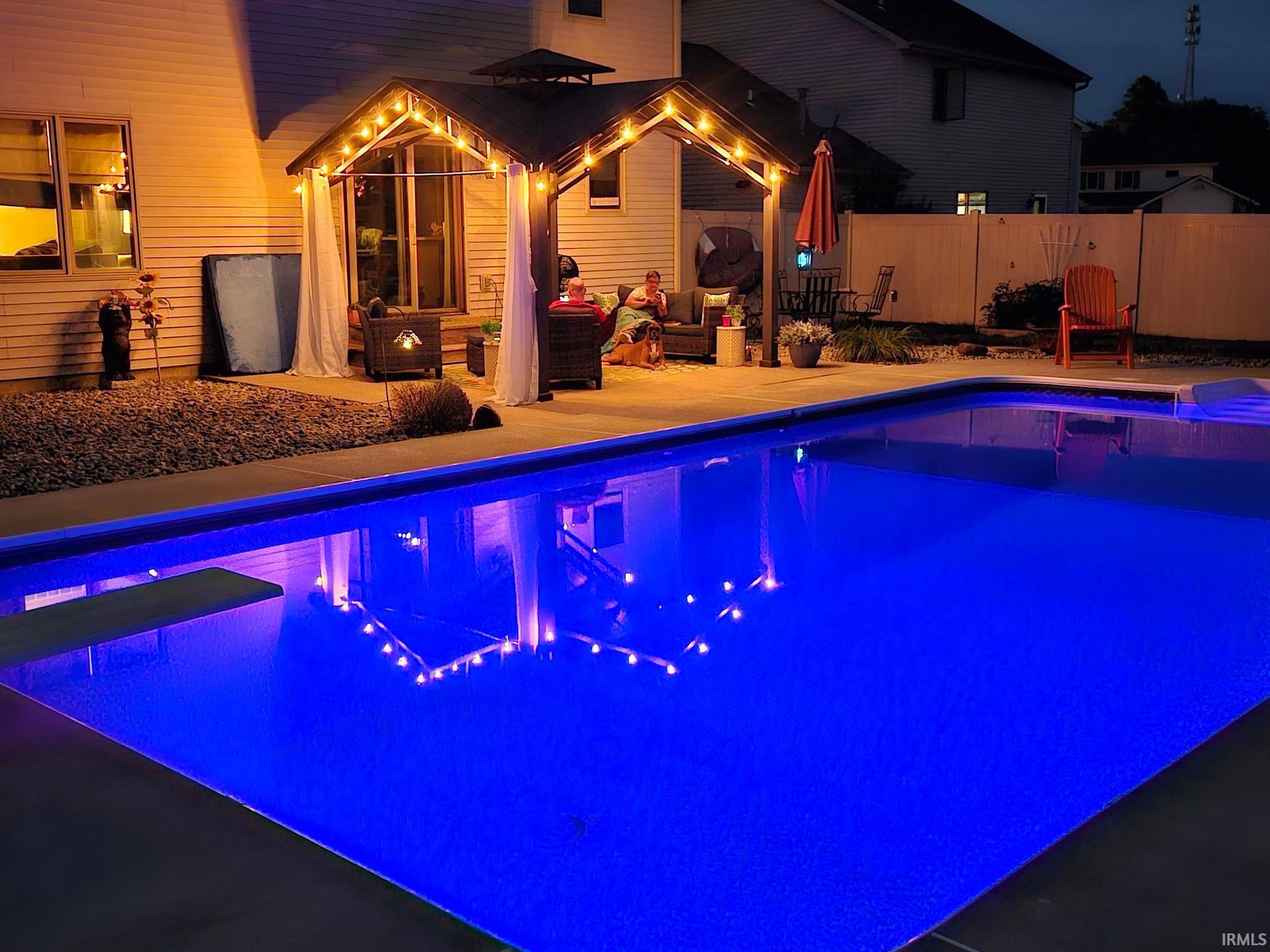Illuminated swimming pool with a diving board, surrounded by concrete decking and pea gravel landscaping