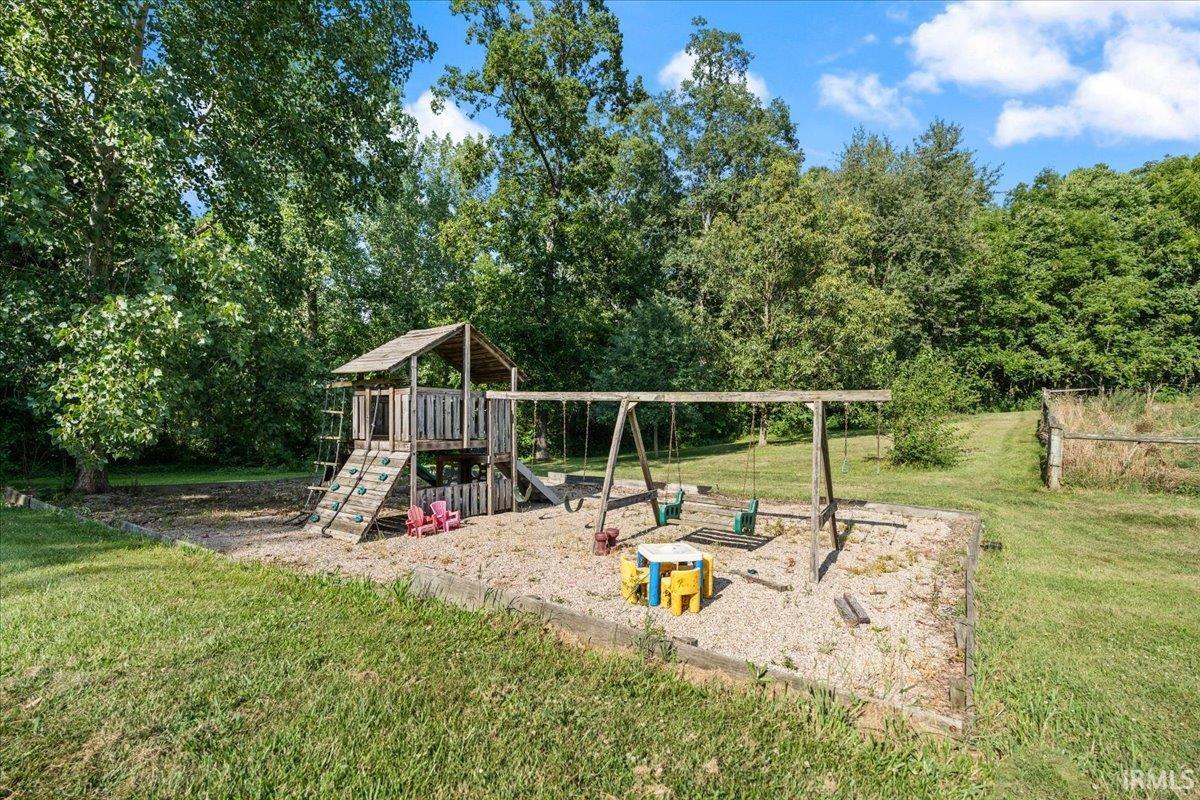 Expansive yard featuring a dedicated play area with a wooden playset, swings, and a gravel base, bordered by mature trees and a grassy field