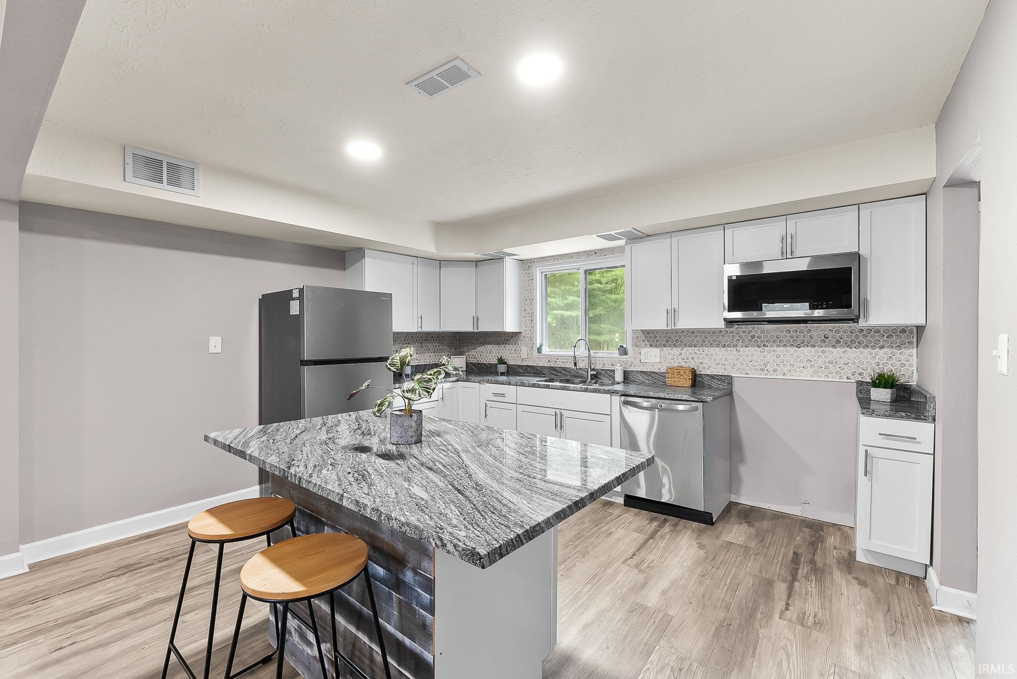 Kitchen featuring an island with stone-finish countertop, stainless steel appliances, light gray cabinetry, mosaic tile backsplash, and wood-finish flooring