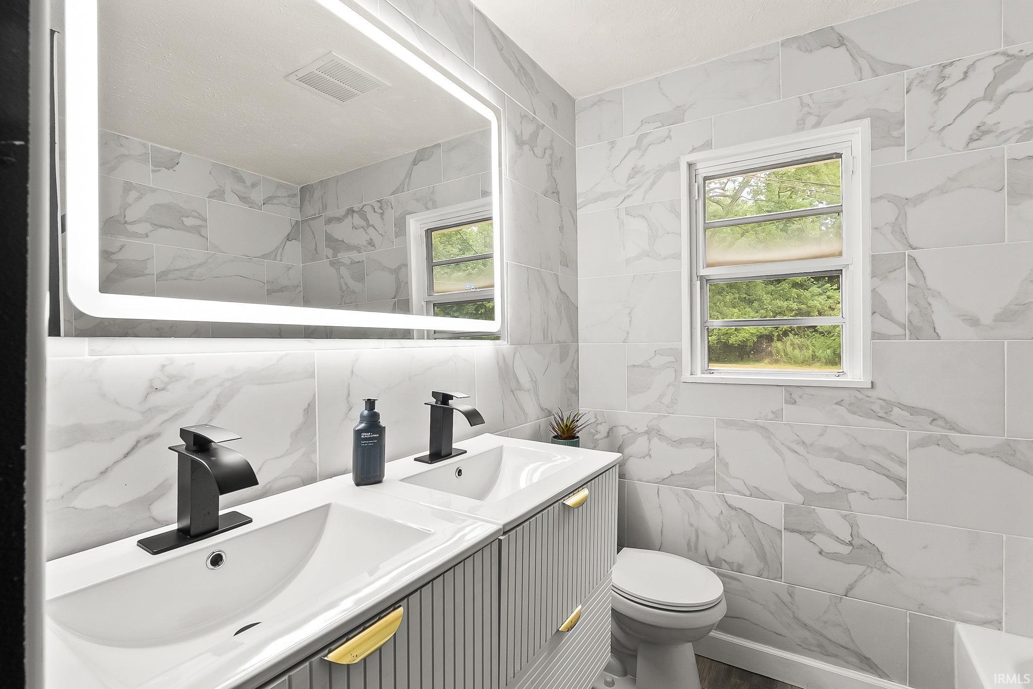 Bathroom featuring a double vanity with matte black faucets, an illuminated mirror, and marble-look wall tiling