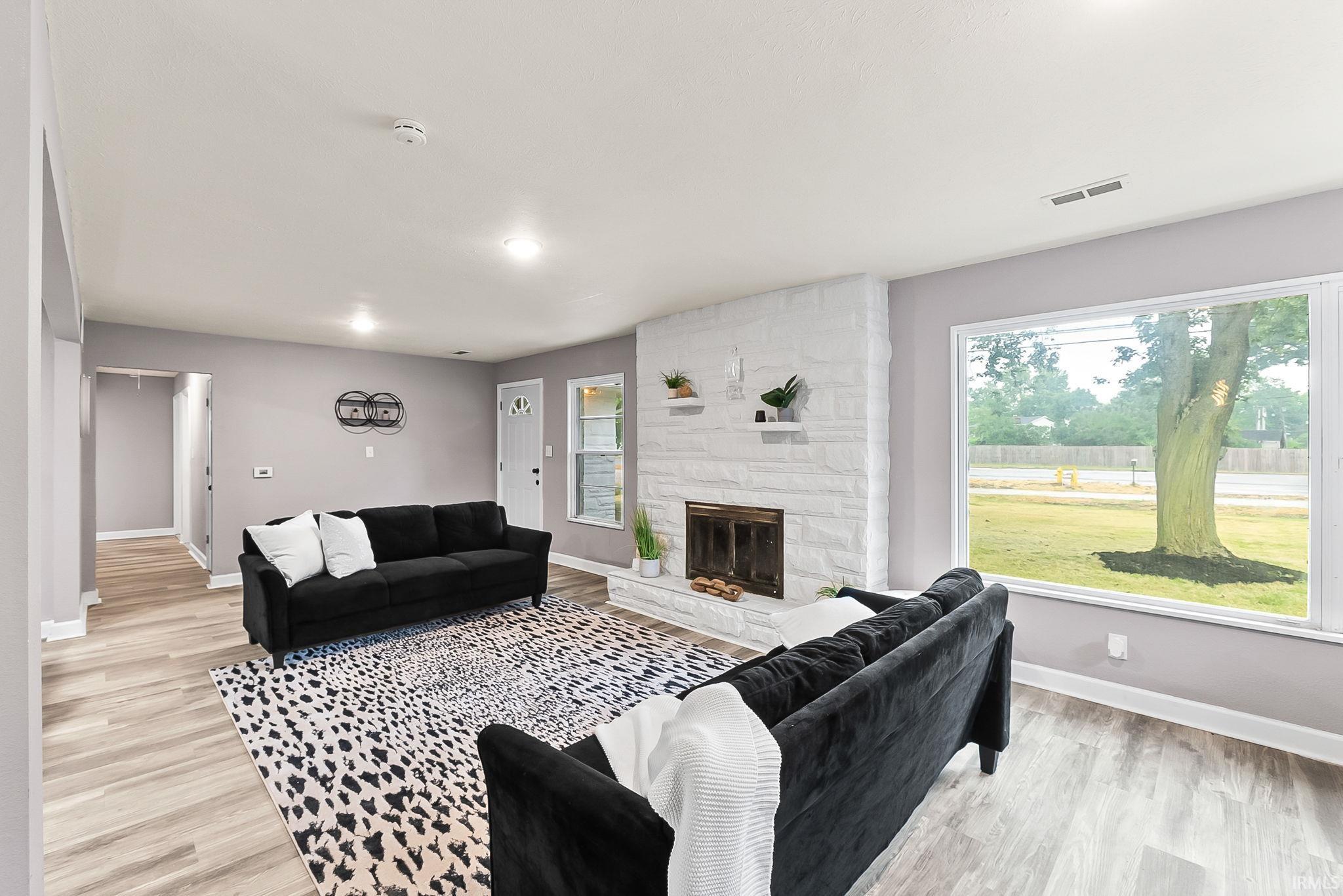 Living room featuring a whitewashed brick fireplace, wood-finish flooring, a large picture window, recessed lighting, and a white paneled entry door