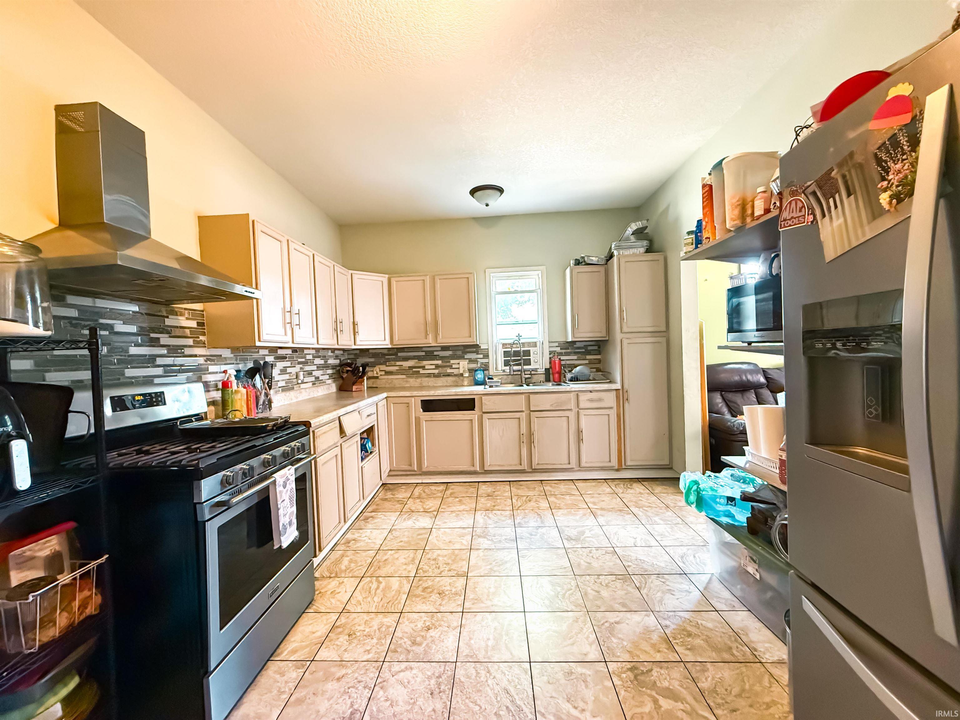 Kitchen featuring tile flooring, light-toned cabinetry, an integrated stainless steel range hood, a patterned tile backsplash, and a double basin sink with a window above