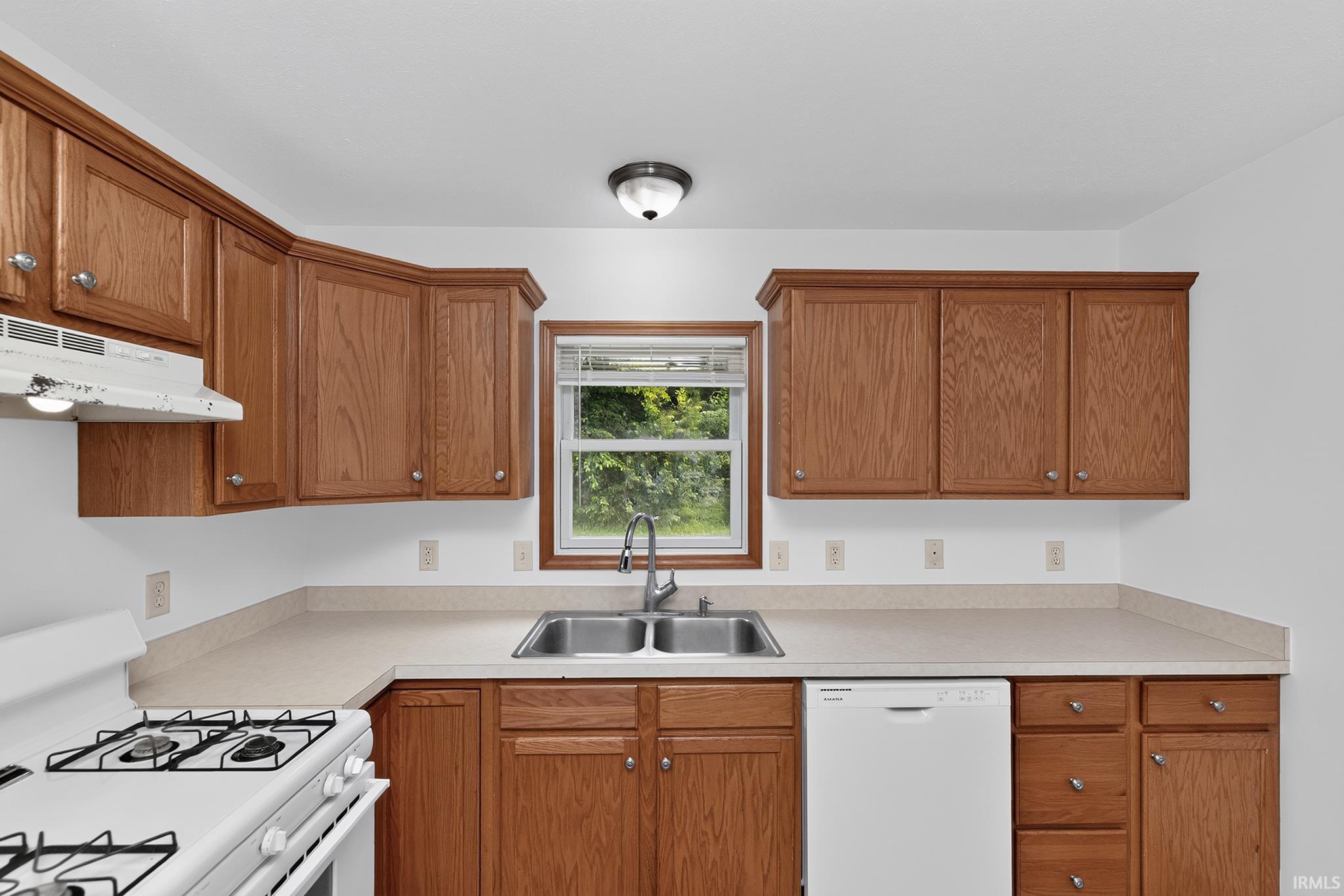 Kitchen featuring wood-finish cabinetry, light-toned countertops, a double basin stainless steel sink, a window with blinds, and white appliances including a range and dishwasher