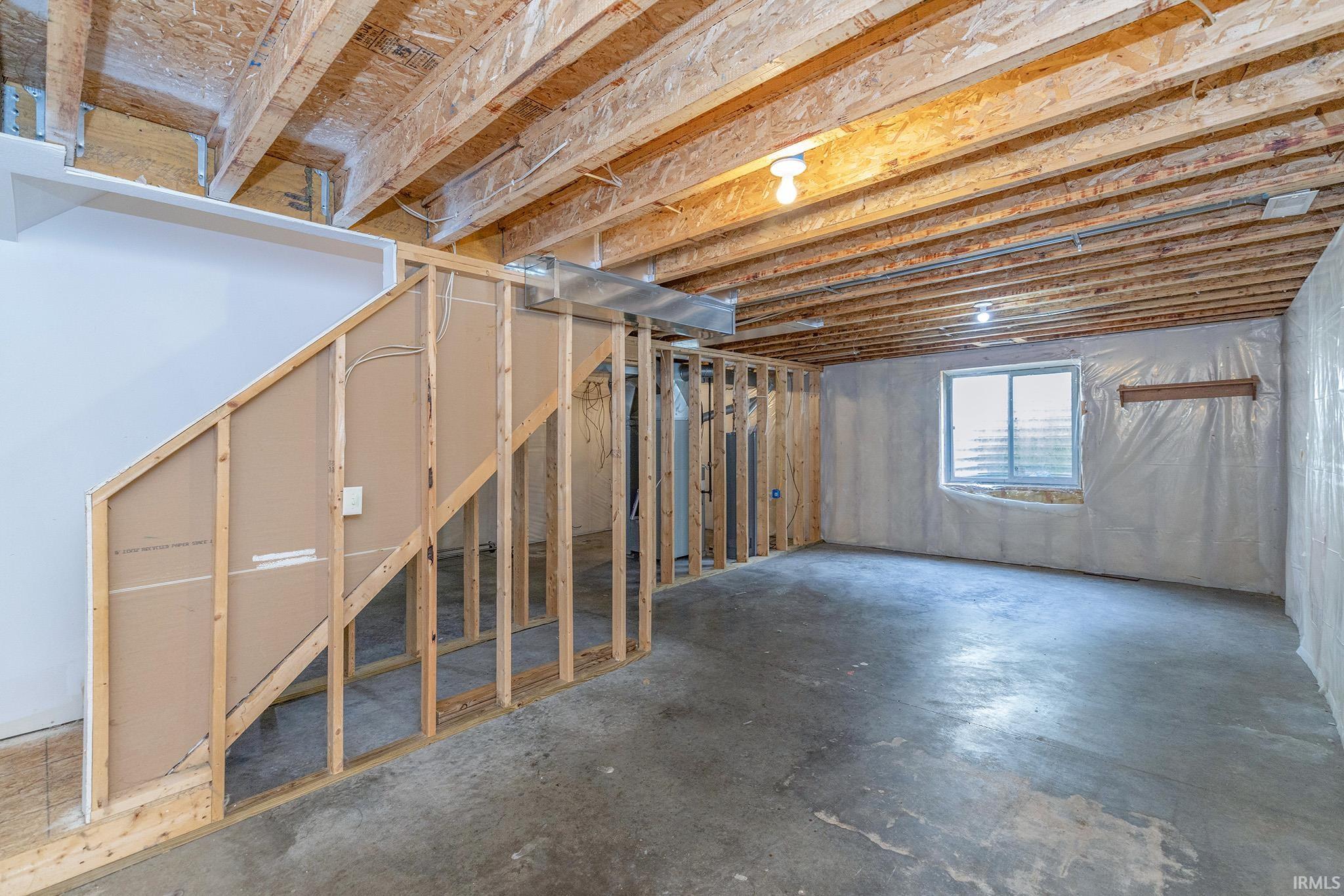 Unfinished basement space featuring exposed wood joists, framed wall sections, concrete flooring, a window, and overhead lighting fixtures