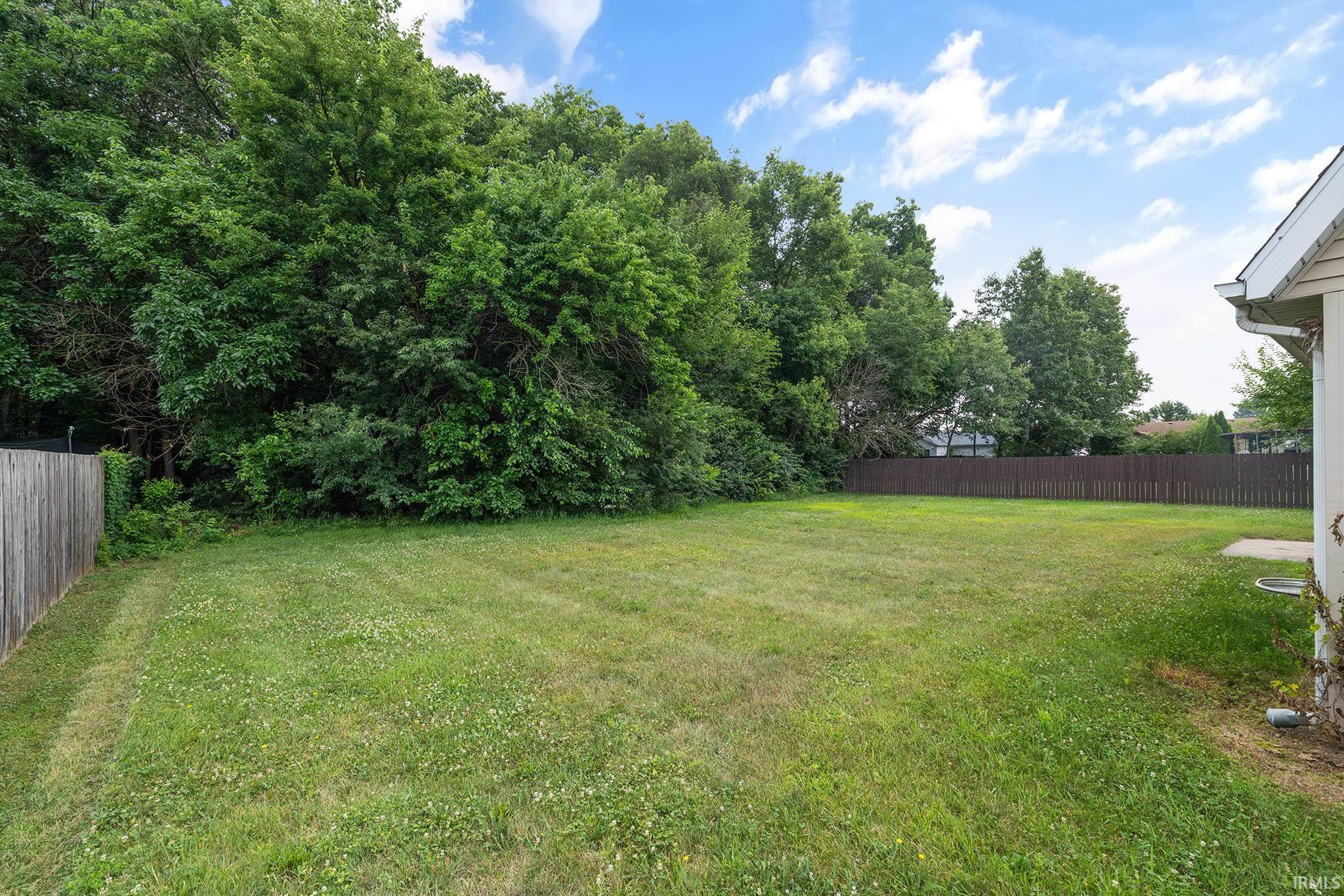Expansive lawn area featuring a wooden privacy fence, mature trees bordering the property, and a concrete patio section