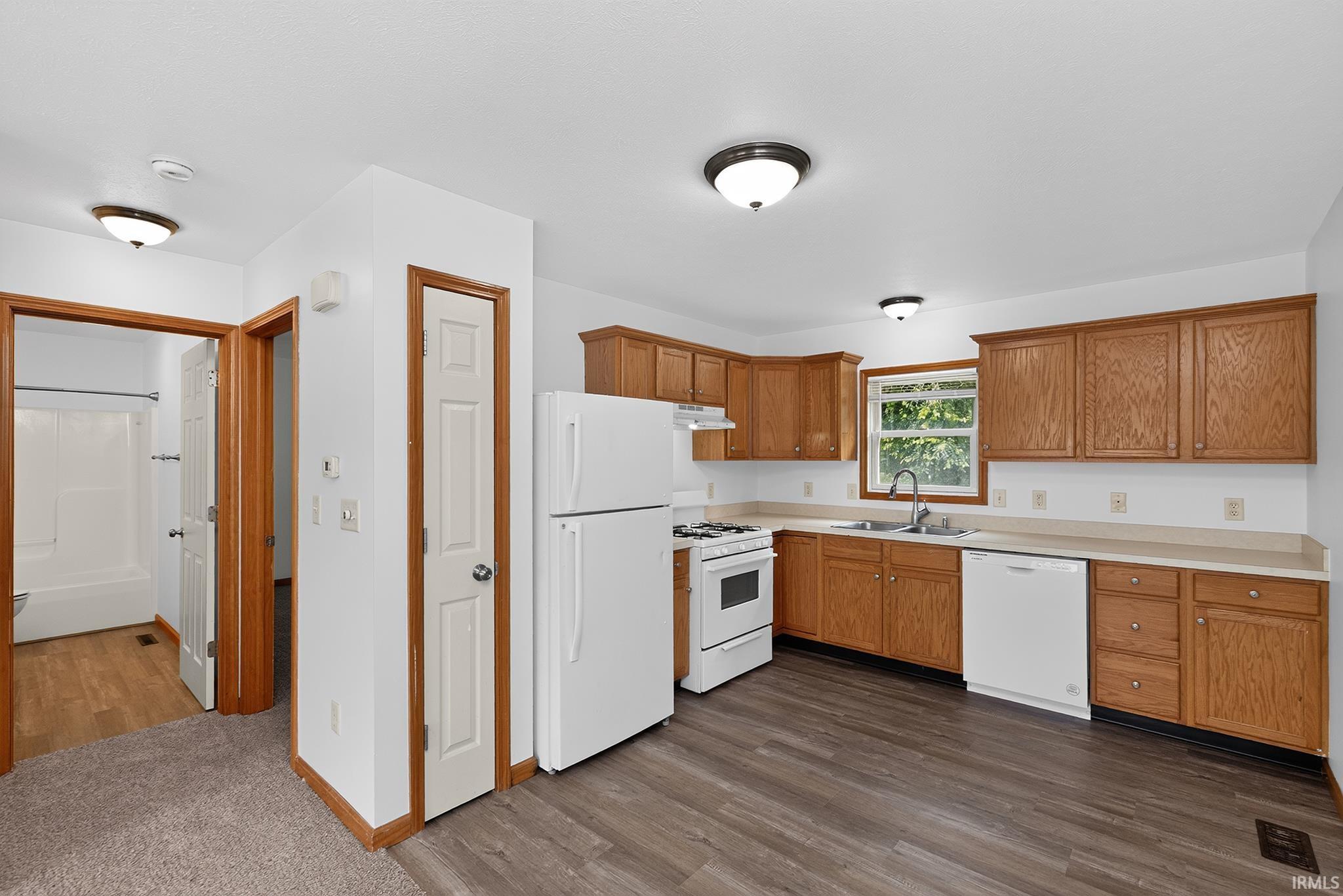 Open-concept kitchen featuring wood-finish flooring, light wood cabinetry, white appliances, and a window above the sink