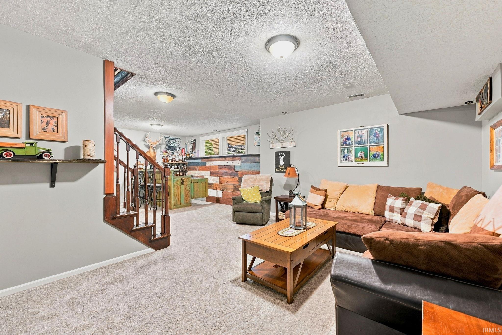 Finished basement featuring light gray walls, light carpet flooring, a dark wood-stained staircase with white risers, and a built-in bar area with a wood-paneled facade