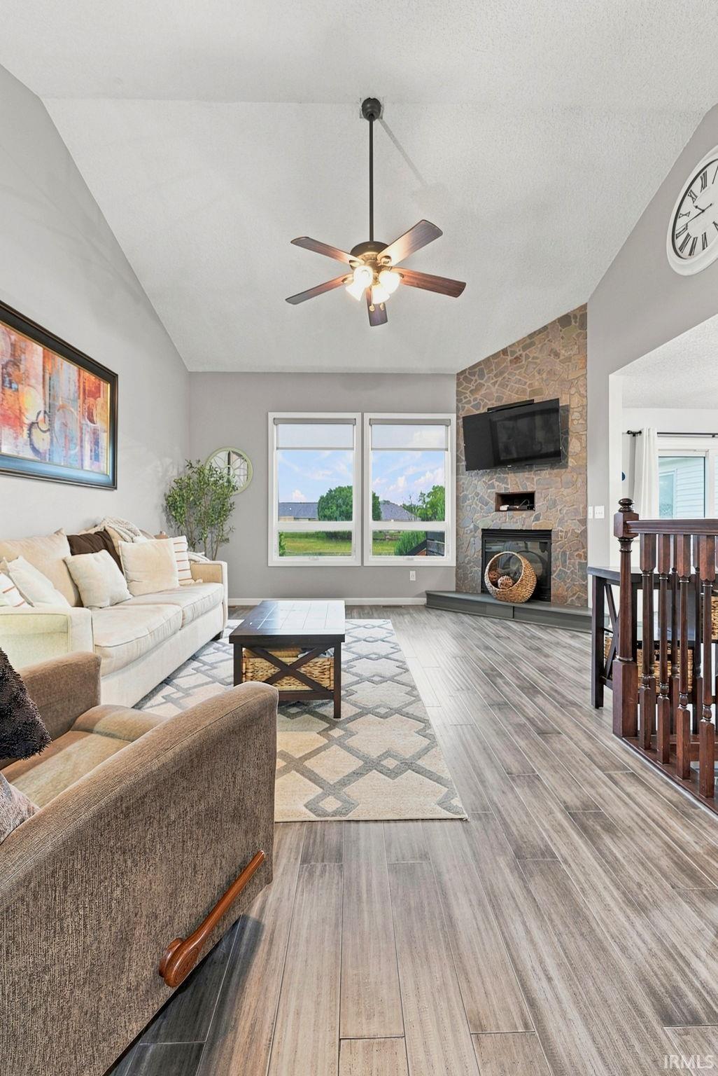 Spacious living area featuring high vaulted ceilings, light wood-finish flooring, a stone fireplace, and a ceiling fan with integrated lighting
