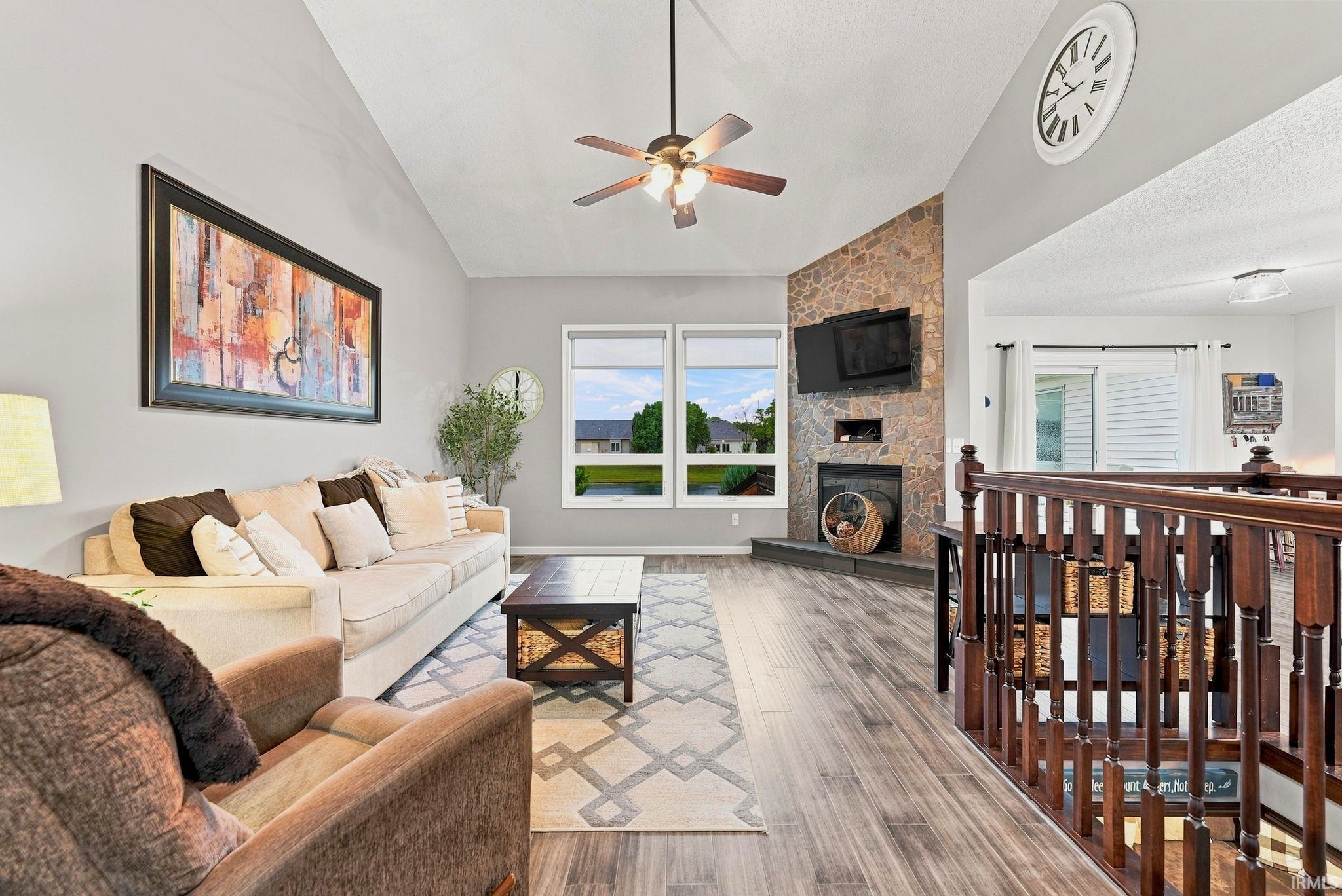 Vaulted ceilings enhance this living space featuring wood-finish flooring, a stone-faced fireplace with a hearth, and a ceiling fan