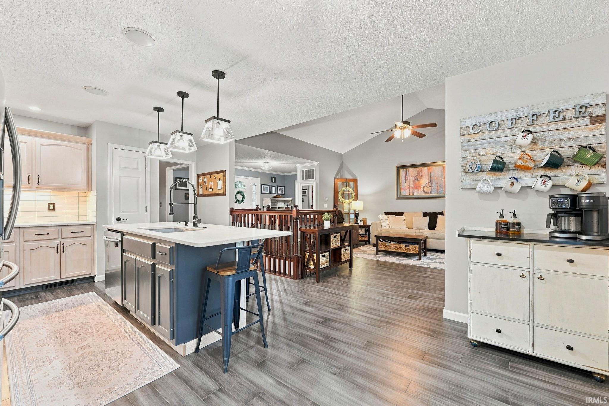 Open-concept kitchen featuring a central island with integrated sink, pendant lighting, shaker-style cabinetry, and wood-finish flooring