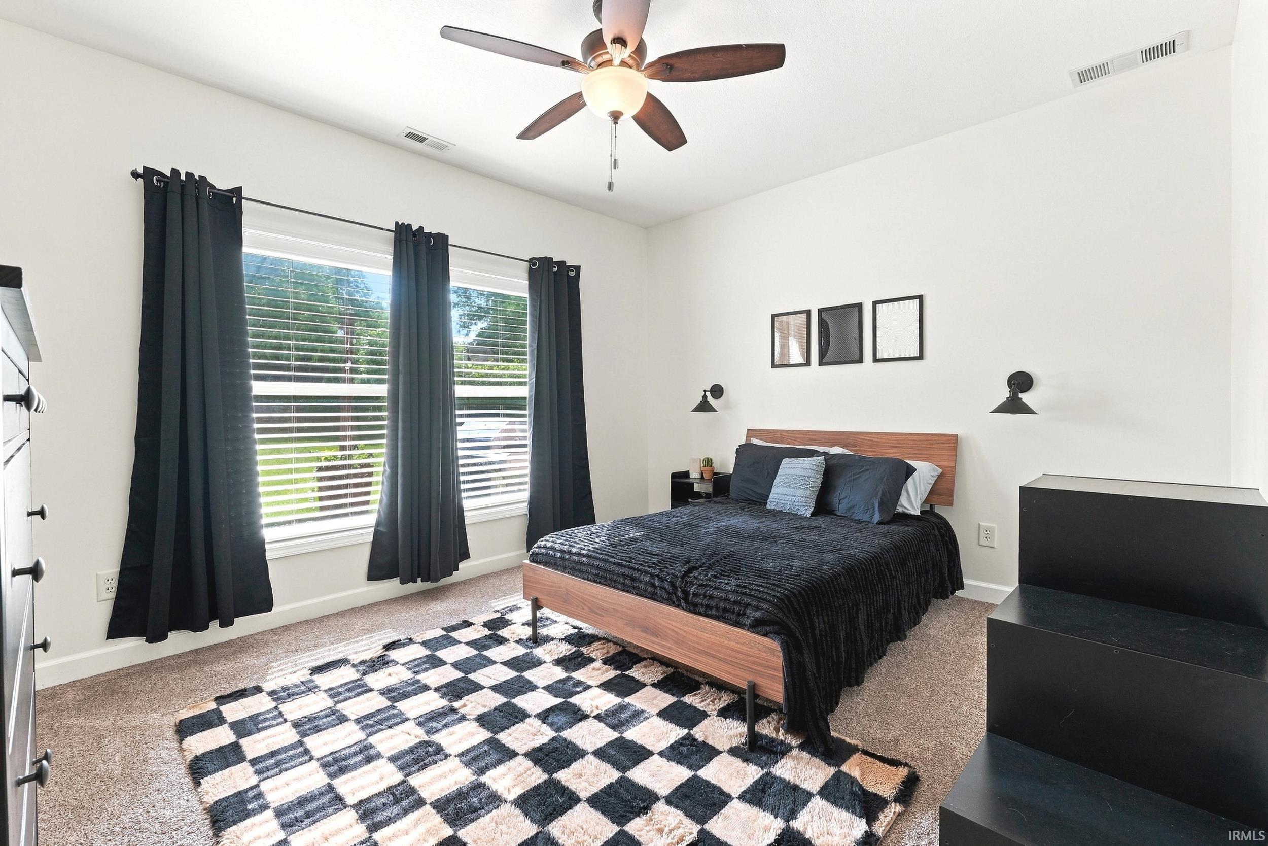 Room featuring a ceiling fan with integrated lighting, three windows with blinds, black wall sconces, and neutral carpeting