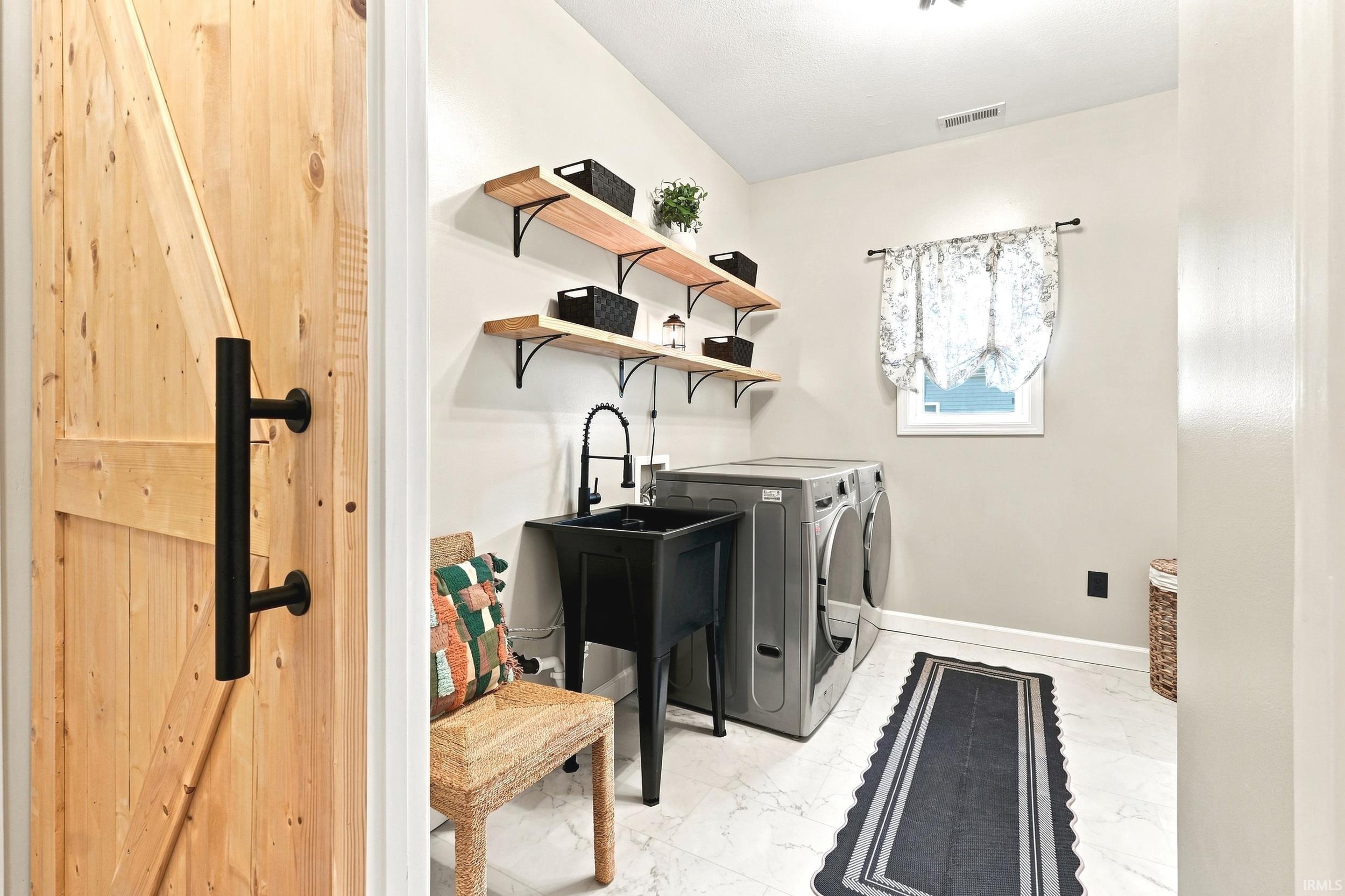 Light wood barn door with black hardware opening into a laundry space featuring white-finish flooring, a utility sink with a matte black faucet, and two wall-mounted wood shelves with black brackets