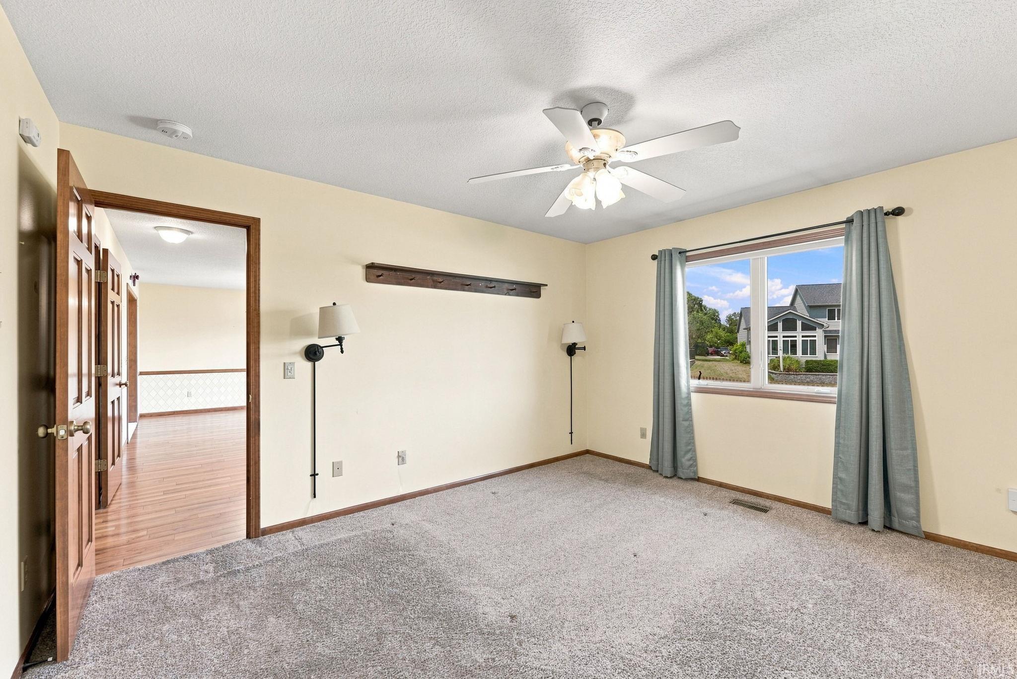 Carpeted room with a ceiling fan, a window with exterior views, and a doorway opening to wood-finish flooring