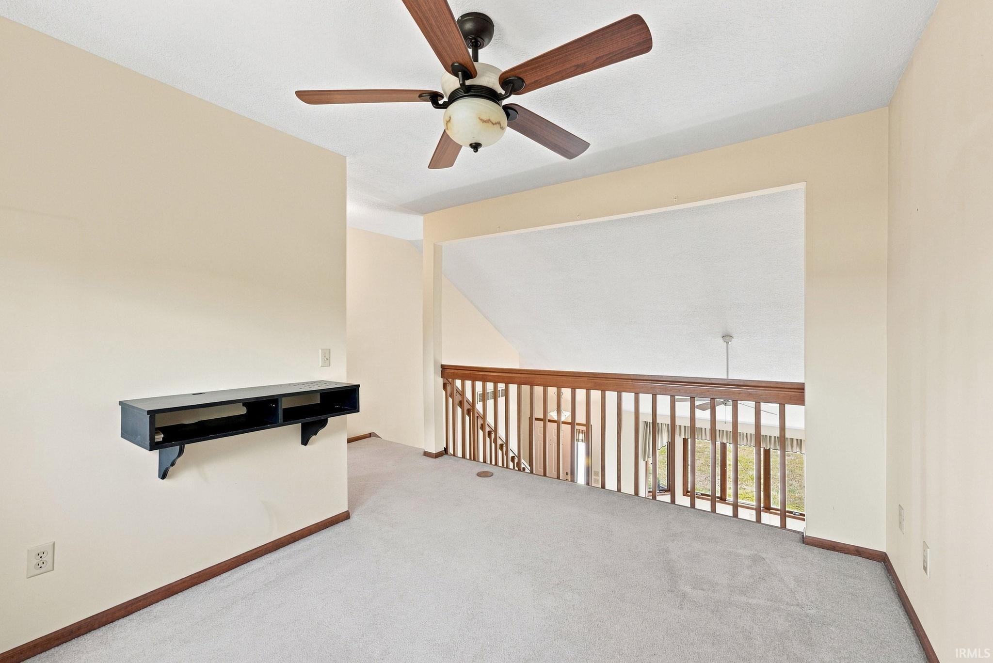 Carpeted loft area featuring a wood-finish railing, a built-in wall shelf, a ceiling fan with integrated lighting, and neutral wall tones