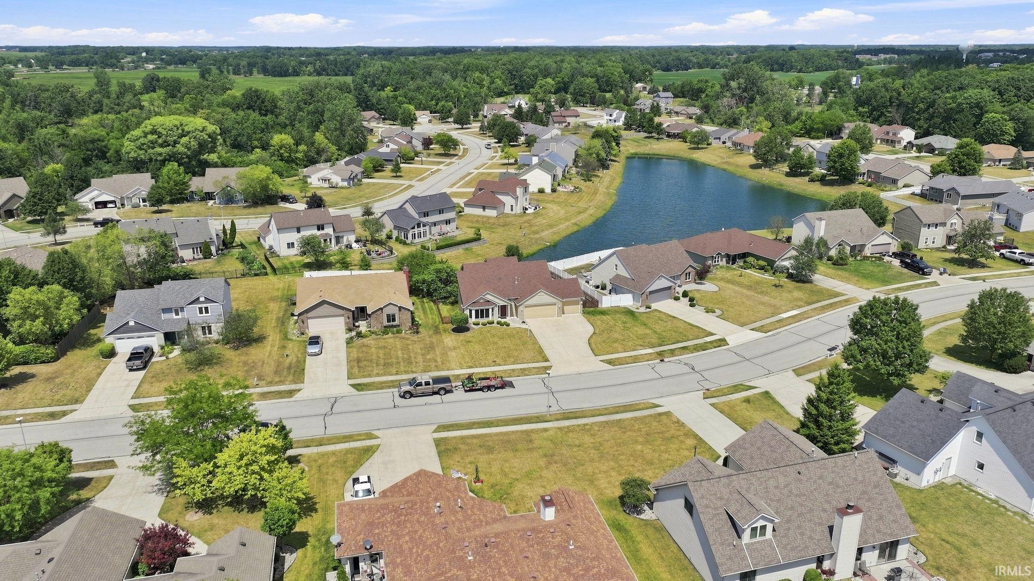 Aerial perspective of a residential neighborhood featuring established homes with varying architectural styles, paved streets, and a central pond