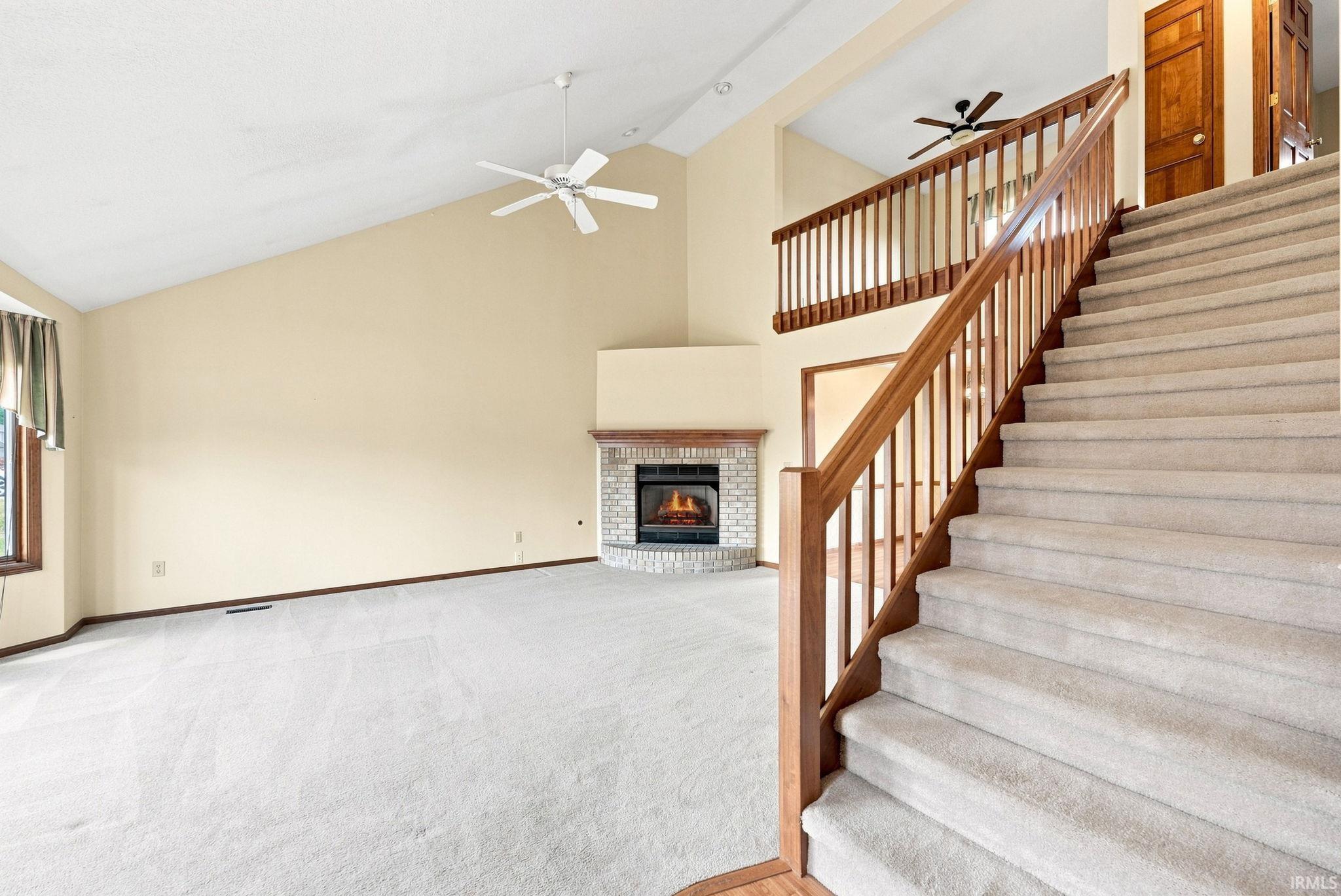 Carpeted staircase with wood banister and railing, opening to a living area featuring vaulted ceilings, a ceiling fan, and a corner brick fireplace with a wood mantel