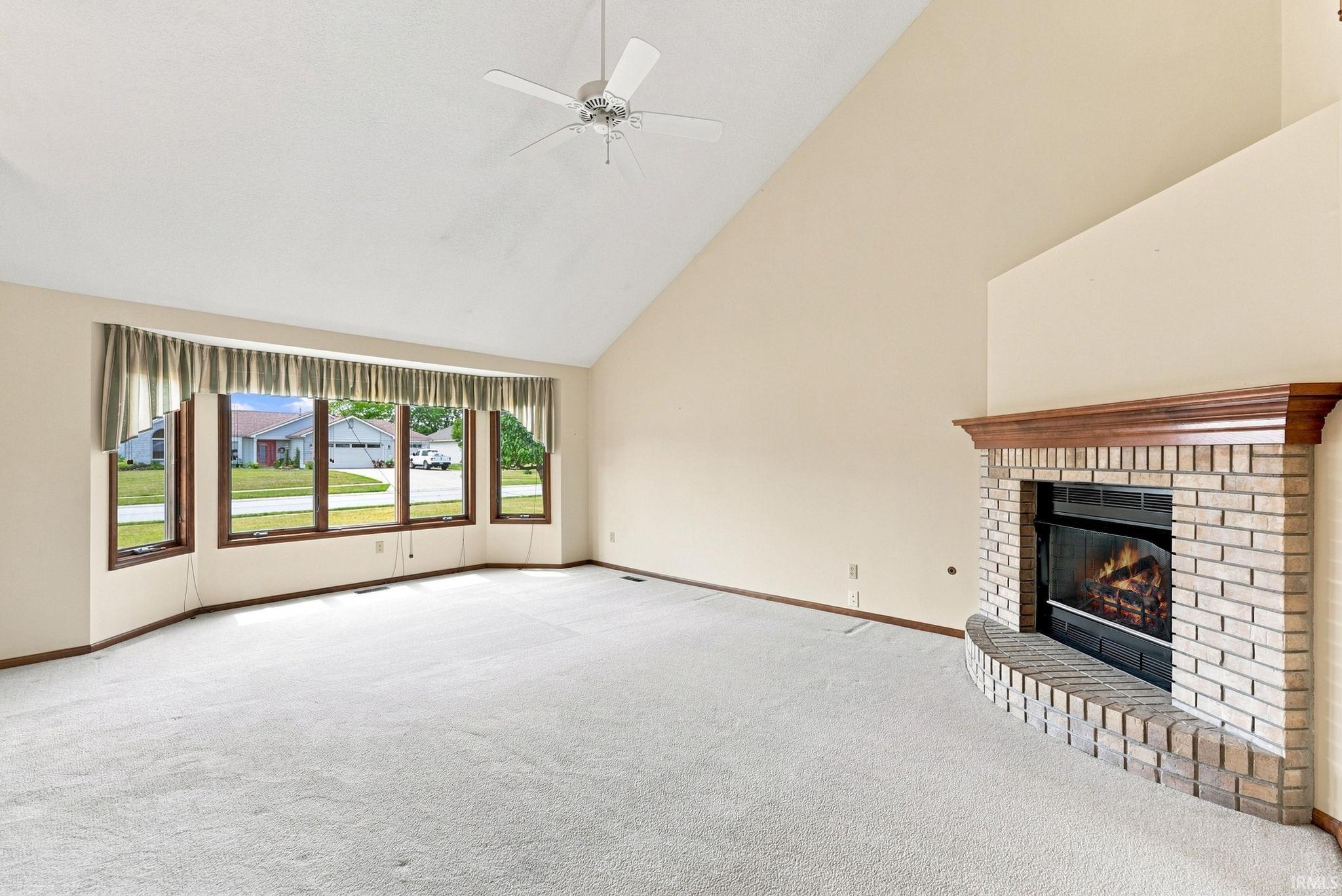 Vaulted ceiling with a ceiling fan, bay window, light-toned carpet flooring, brick fireplace with a wood mantle, and neutral wall tones