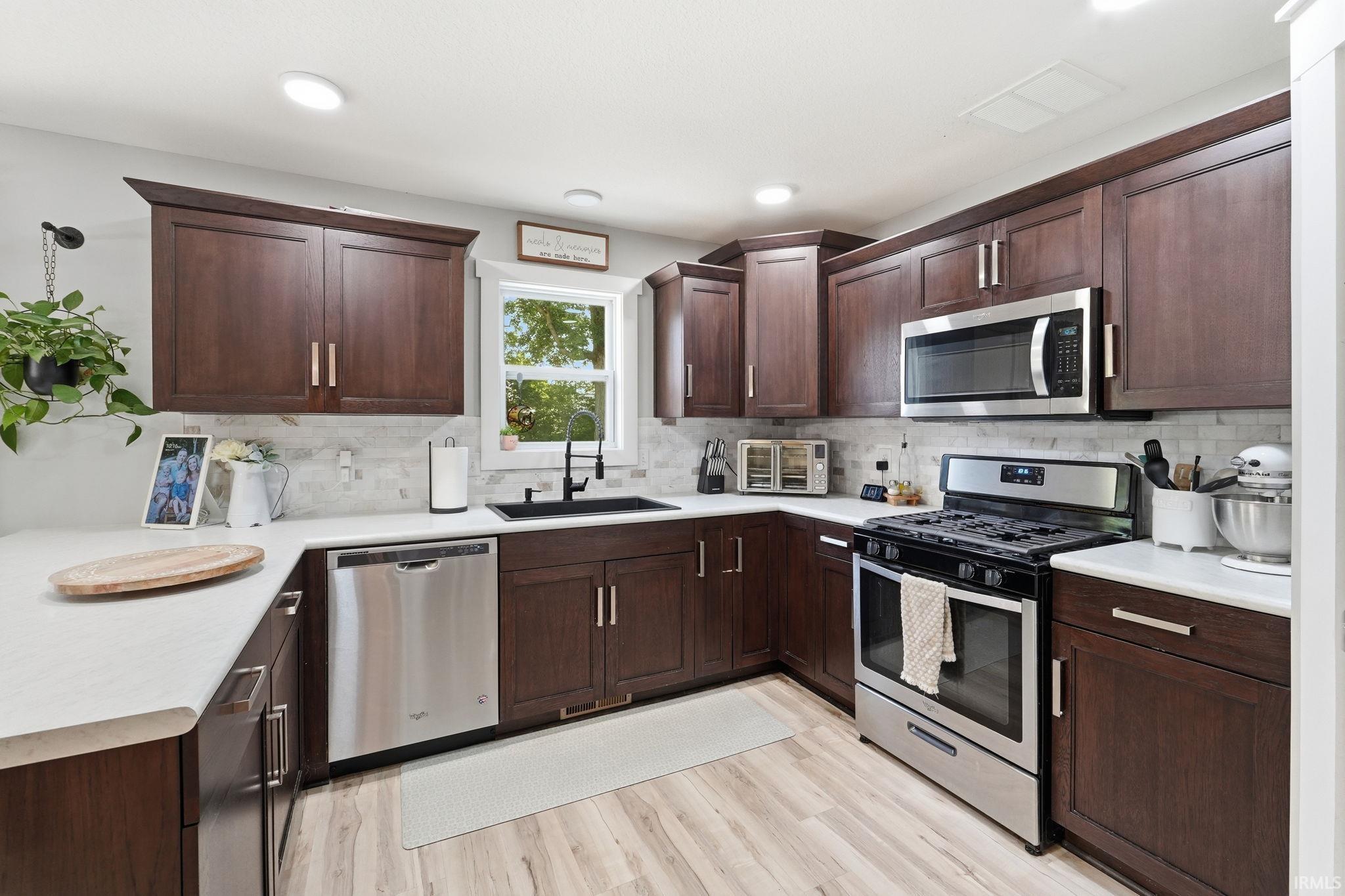 Modern kitchen featuring dark wood cabinetry, light countertops, stainless steel appliances, a tile backsplash, and light wood-finish flooring