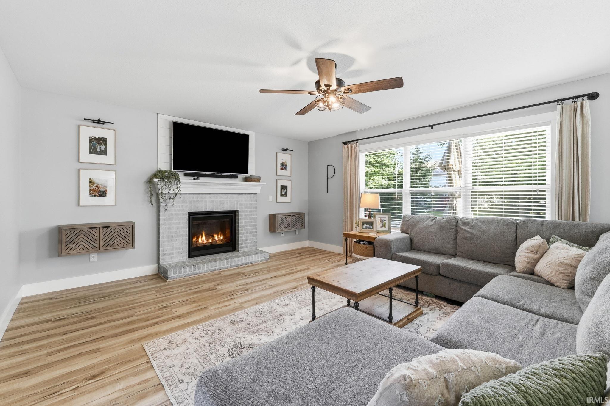 Light-filled living area featuring wood-finish flooring, a painted brick fireplace with a mantle, a large window with blinds, and a ceiling fan with integrated lighting