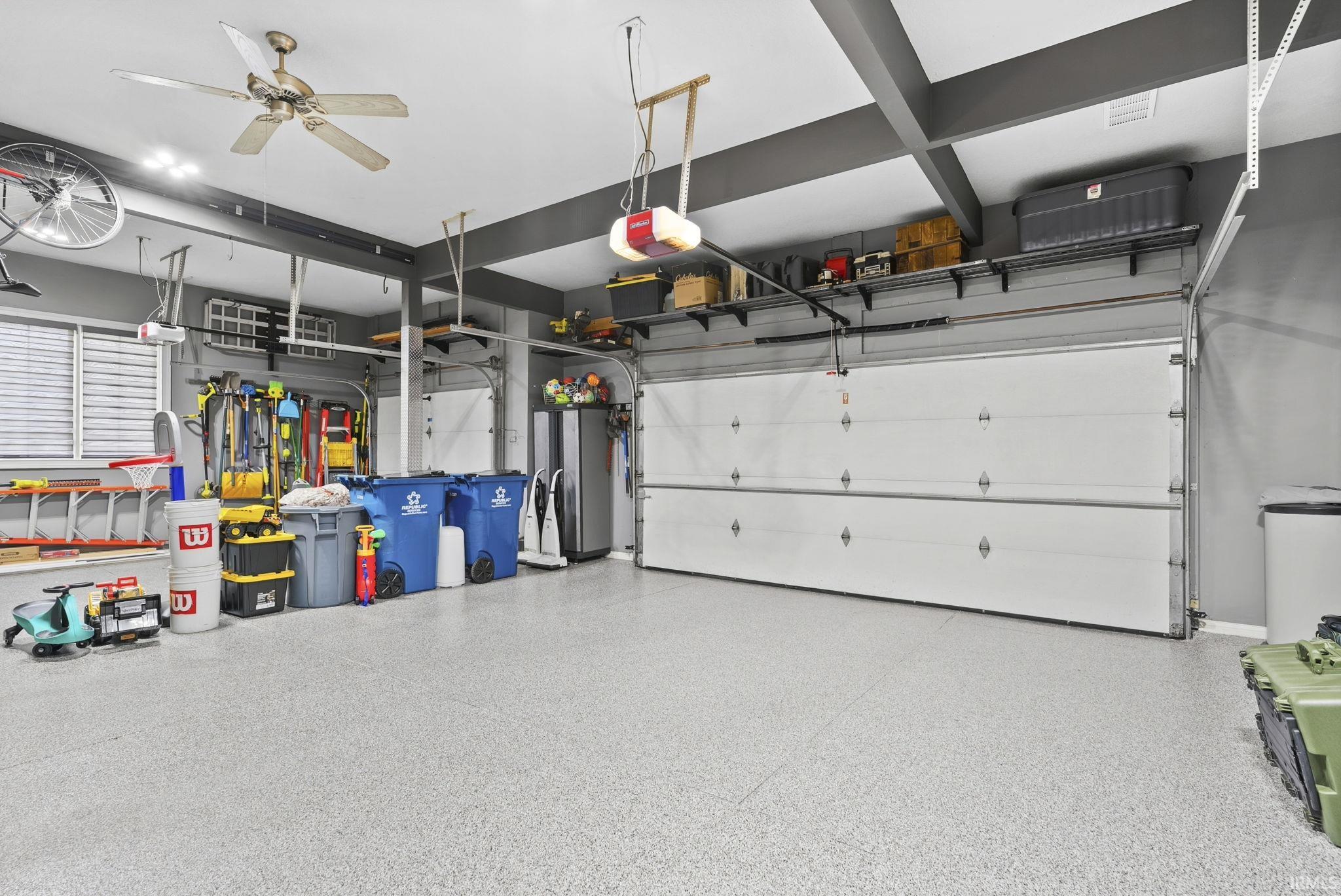 Garage featuring a light-colored epoxy floor, two overhead garage doors, exposed ceiling beams, and a ceiling fan