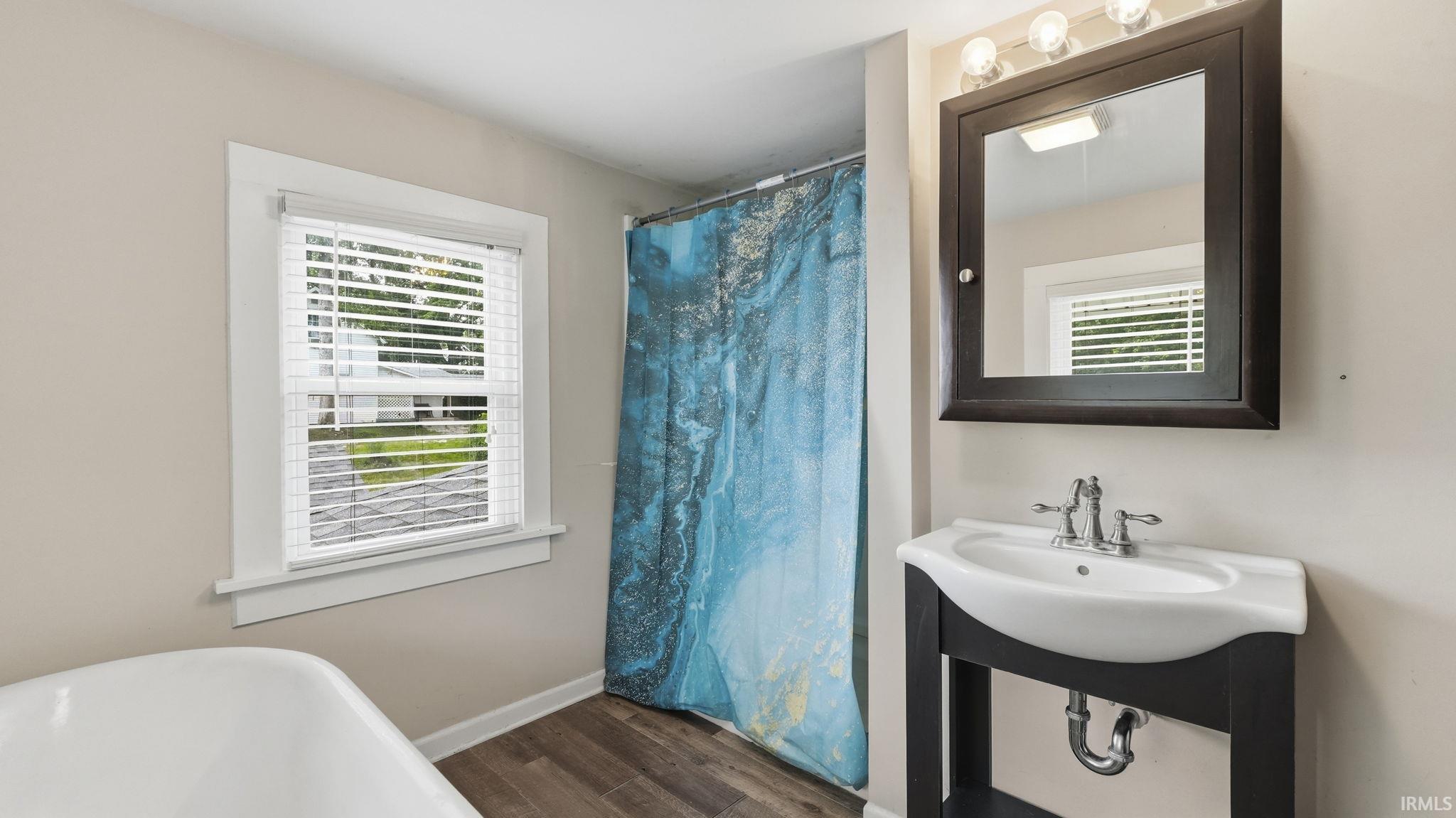 Bathroom featuring a freestanding white bathtub, a single window with white blinds, wood-finish flooring, a dark wood-frame medicine cabinet, and a pedestal sink with a chrome faucet