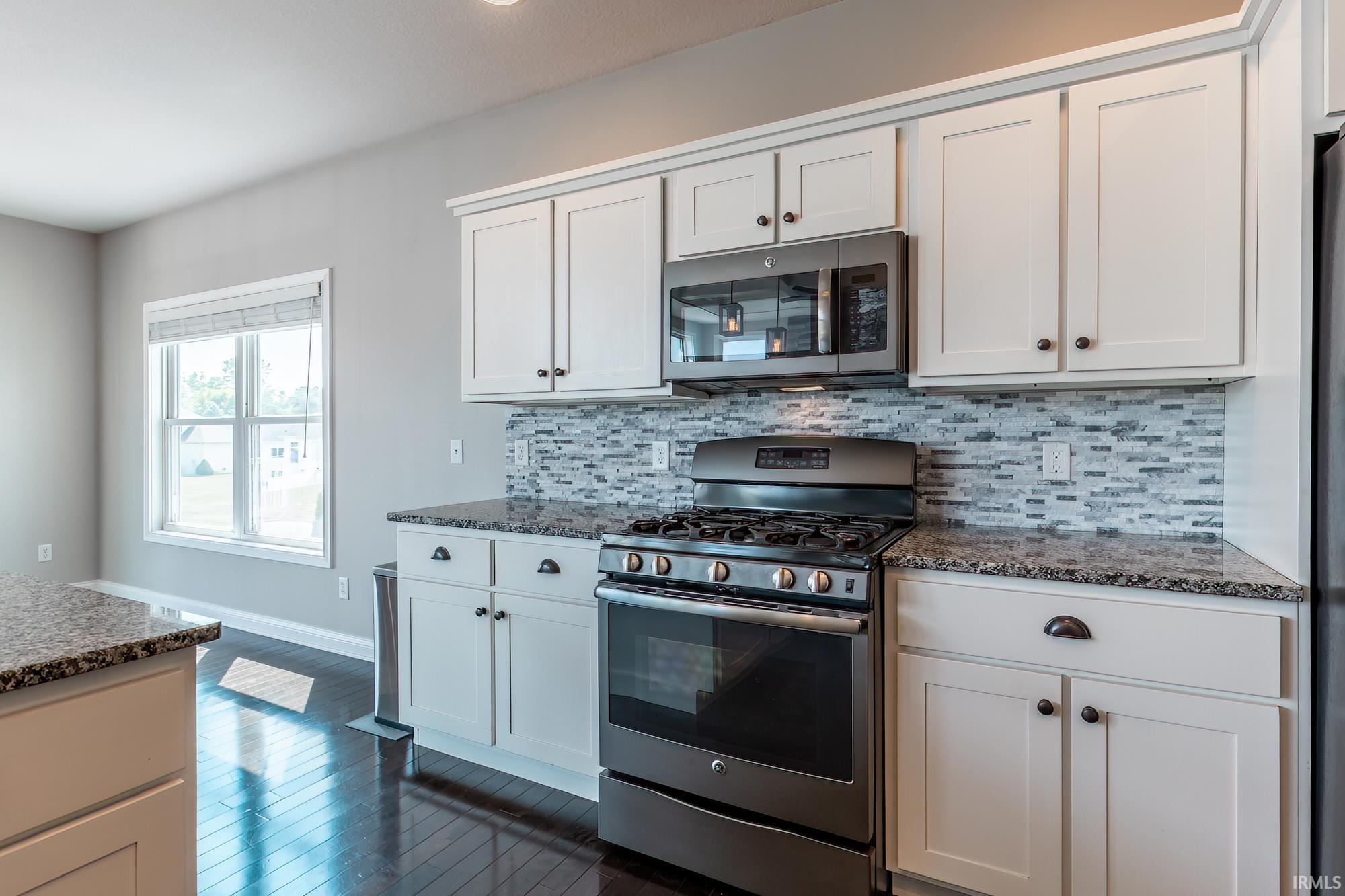Kitchen featuring white shaker-style cabinetry, stainless steel appliance suite, patterned tile backsplash, dark wood-finish flooring, and light-toned countertops