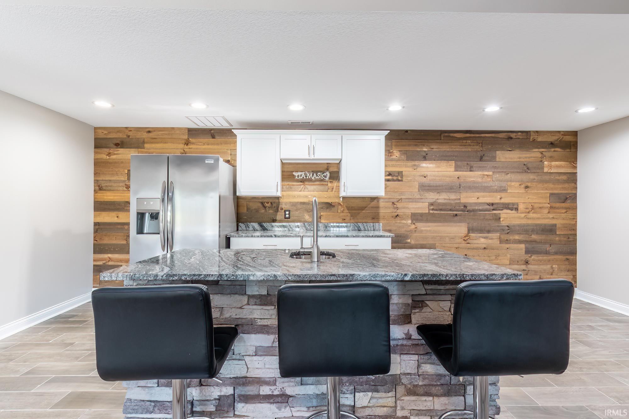 Kitchen featuring a stacked stone island with granite countertop, stainless steel refrigerator, wood plank accent wall, white cabinetry, and recessed lighting