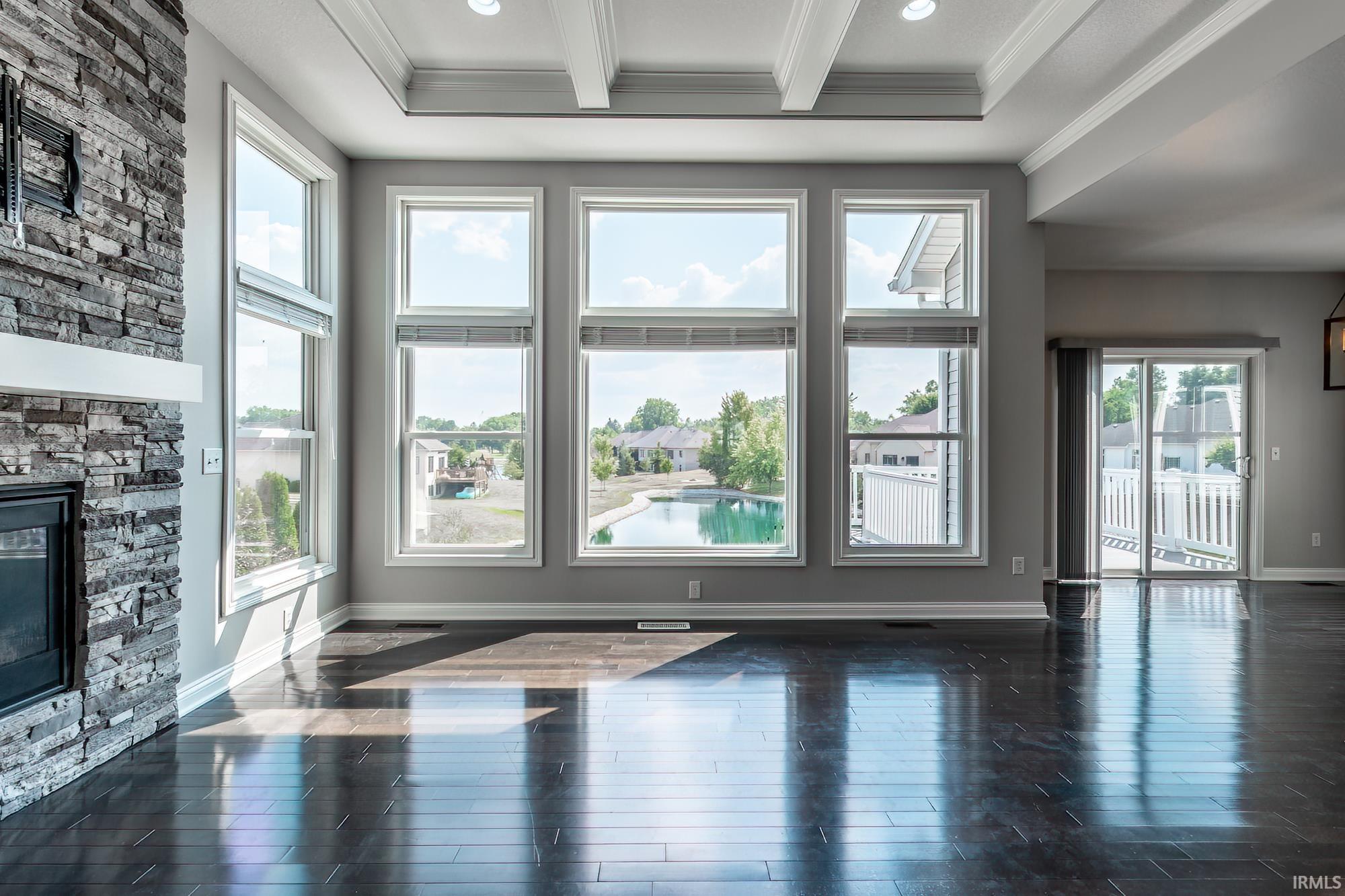 Bright and spacious great room showcasing floor-to-ceiling multi-pane windows with expansive natural light, dark wood-finish flooring, coffered ceiling detail, and a striking stacked stone fireplace accent wall