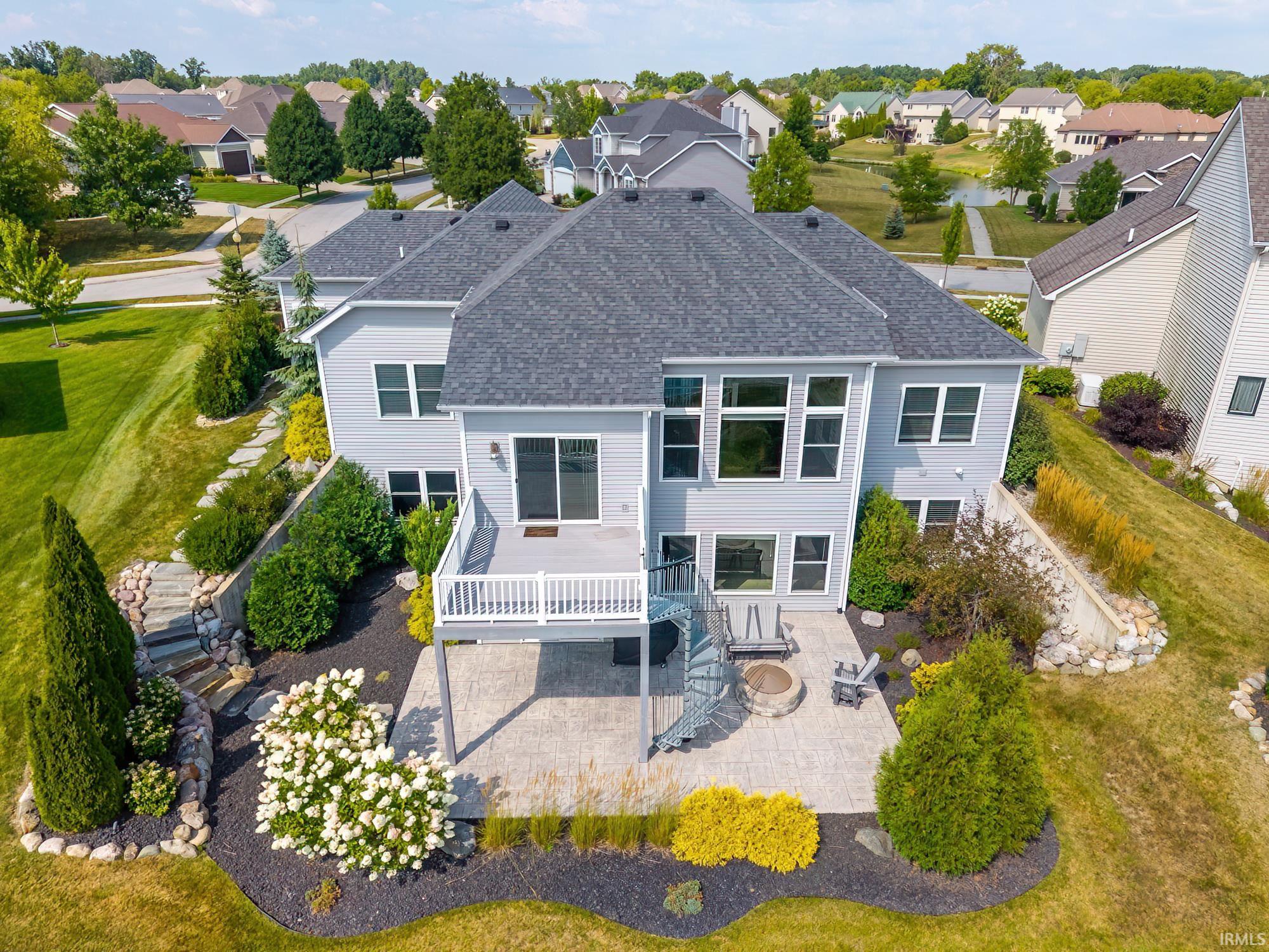 Rear exterior featuring a spacious elevated deck with white railings, a spiral staircase leading to a stone paver patio, multiple windows, and manicured landscaping with mature plantings