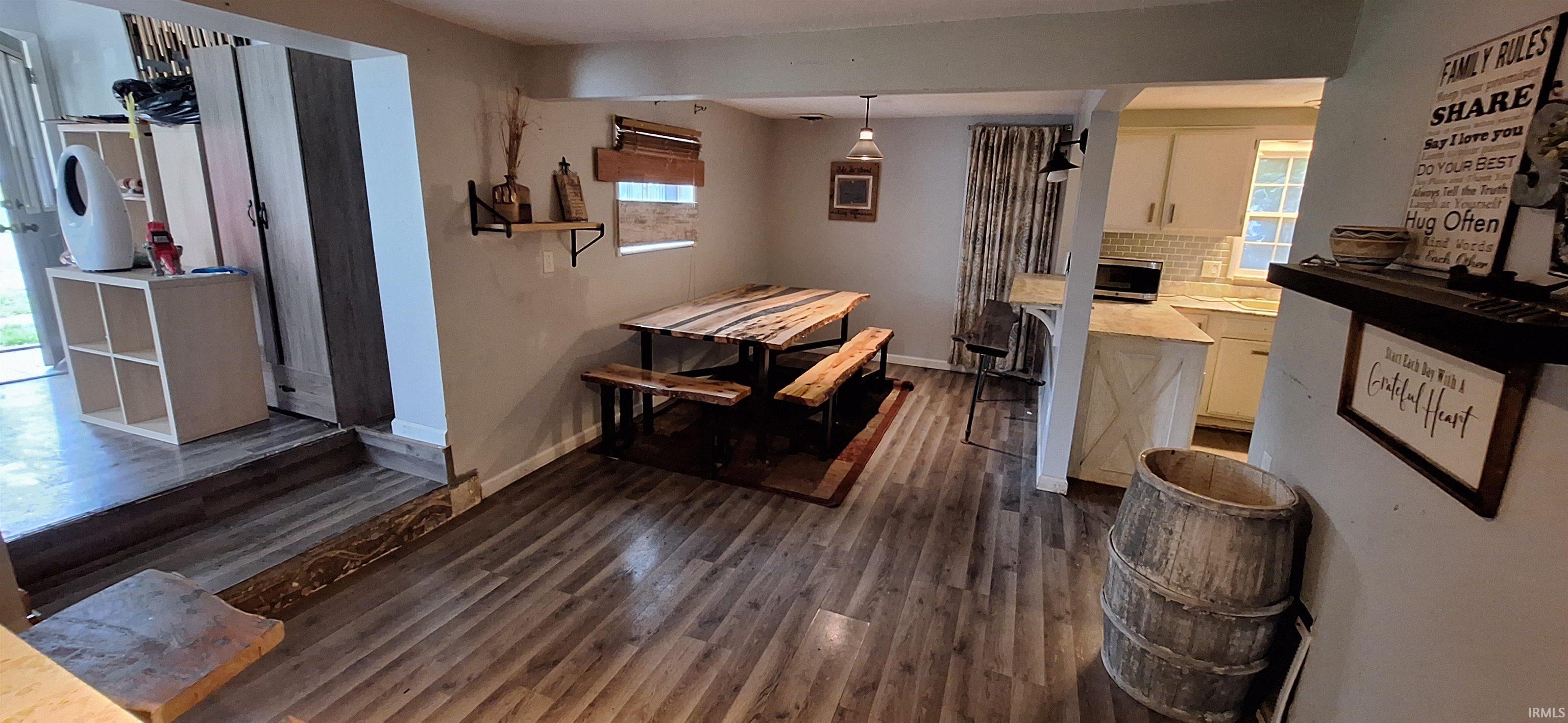 Spacious room featuring wood-finish flooring, a pendant light fixture, and a partial view of the kitchen with white cabinetry and a tiled backsplash