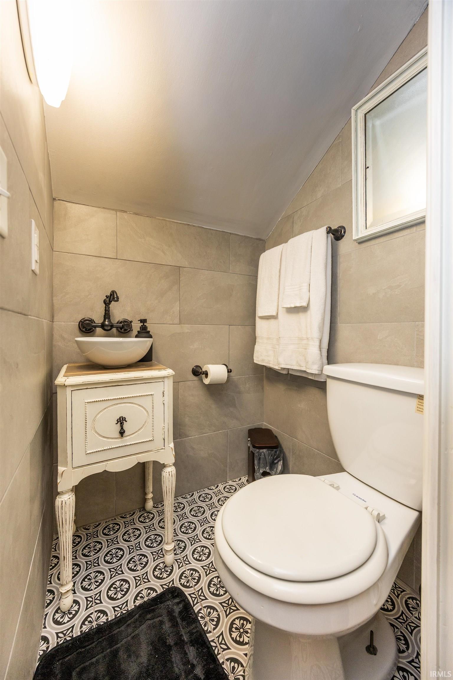 Powder room featuring decorative patterned floor tiles, light-toned wall tiling, a vessel sink with a dark bronze-finish faucet, and a white vanity with distressed detailing
