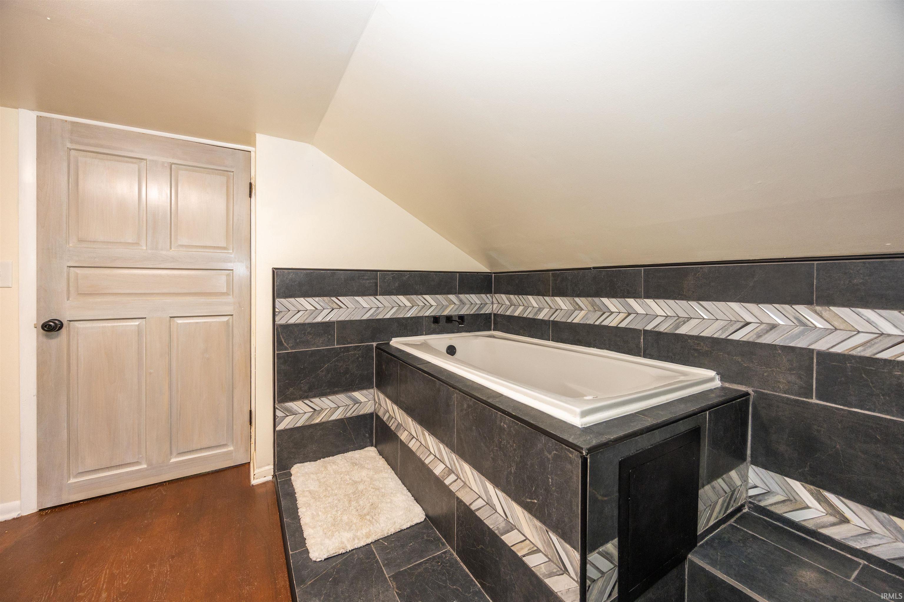 Built-in bathtub surrounded by dark tiled walls with a decorative chevron pattern, dark tiled flooring, and a light-colored paneled door