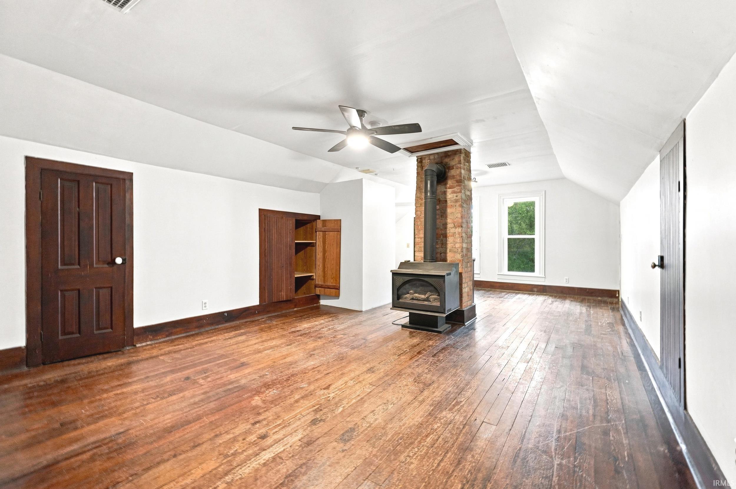 Spacious room featuring wood-finish flooring, a freestanding fireplace with an exposed brick chimney, and a ceiling fan