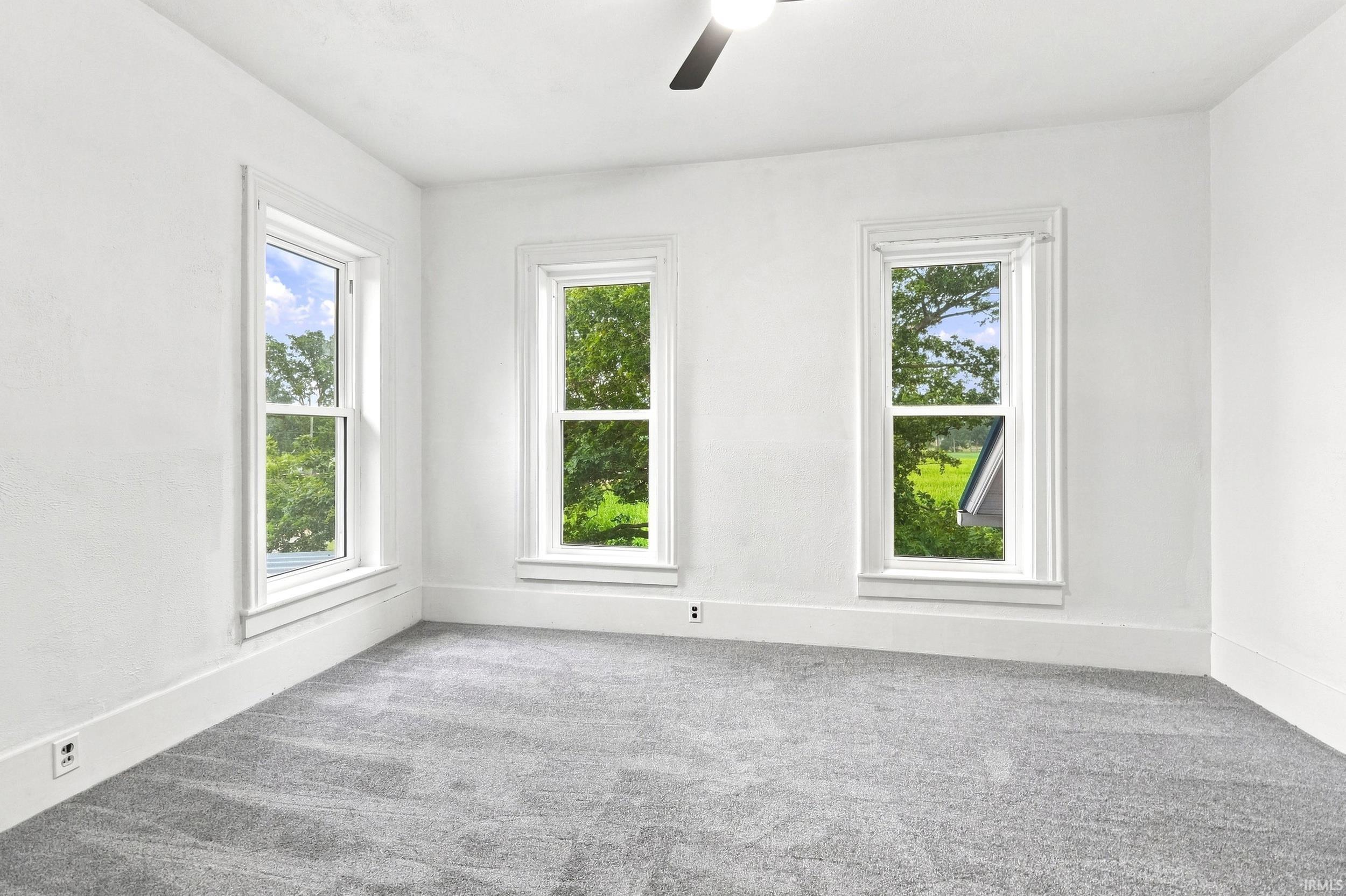 Bright interior space featuring three large windows with white trim, gray carpeting, and a ceiling fan
