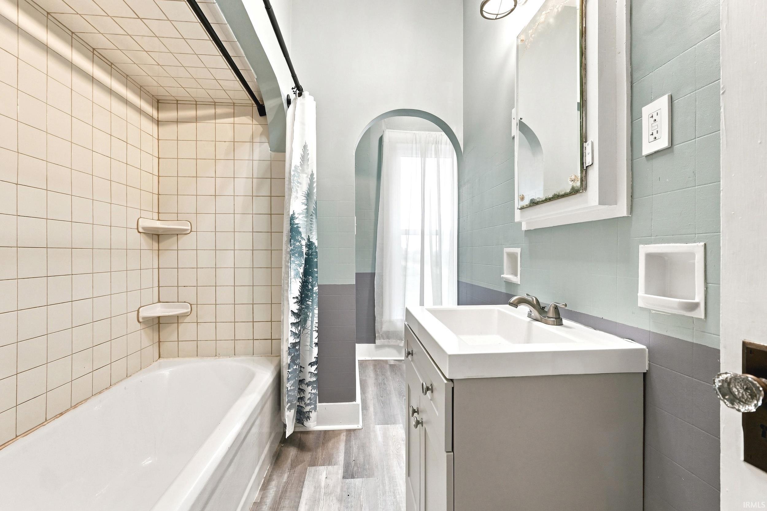 Bathroom featuring a white tub with tiled surround, a vanity with integrated sink, and wood-finish flooring
